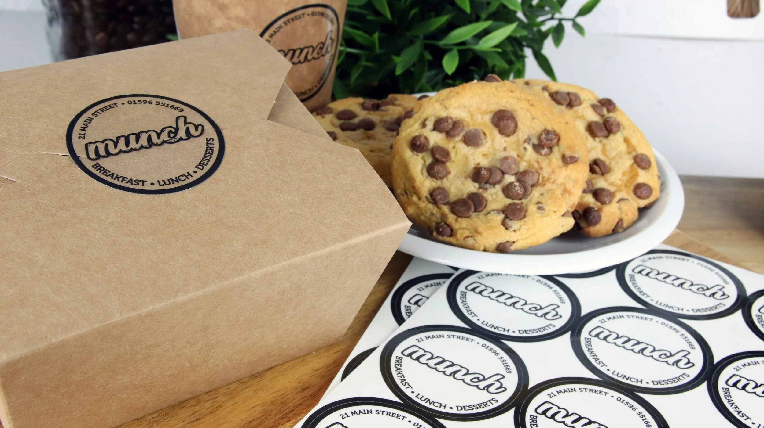A brown paper box with a black circular logo labeled "munch" contains breakfast, lunch, and desserts. Next to it is a white plate with several chocolate chip cookies. There are black and white circular stickers with the same "munch" logo and address on a sheet of paper on a wooden table, with a green plant and a brown paper coffee cup visible in the background.