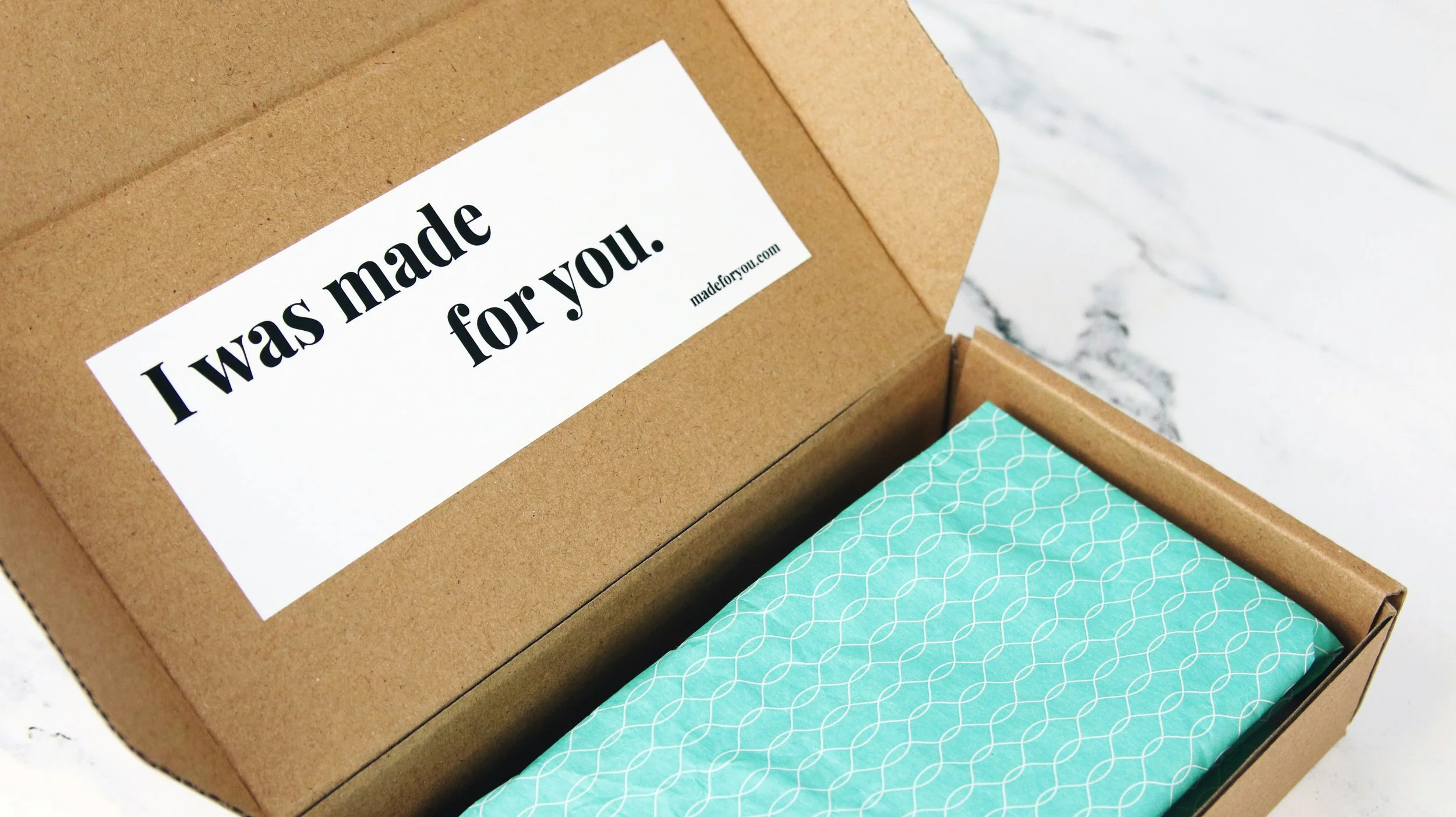 Open brown cardboard box with a white label that reads, 'I was made for you.' Inside, there's turquoise wrapping paper with a geometric pattern. The box rests on a white marble surface.