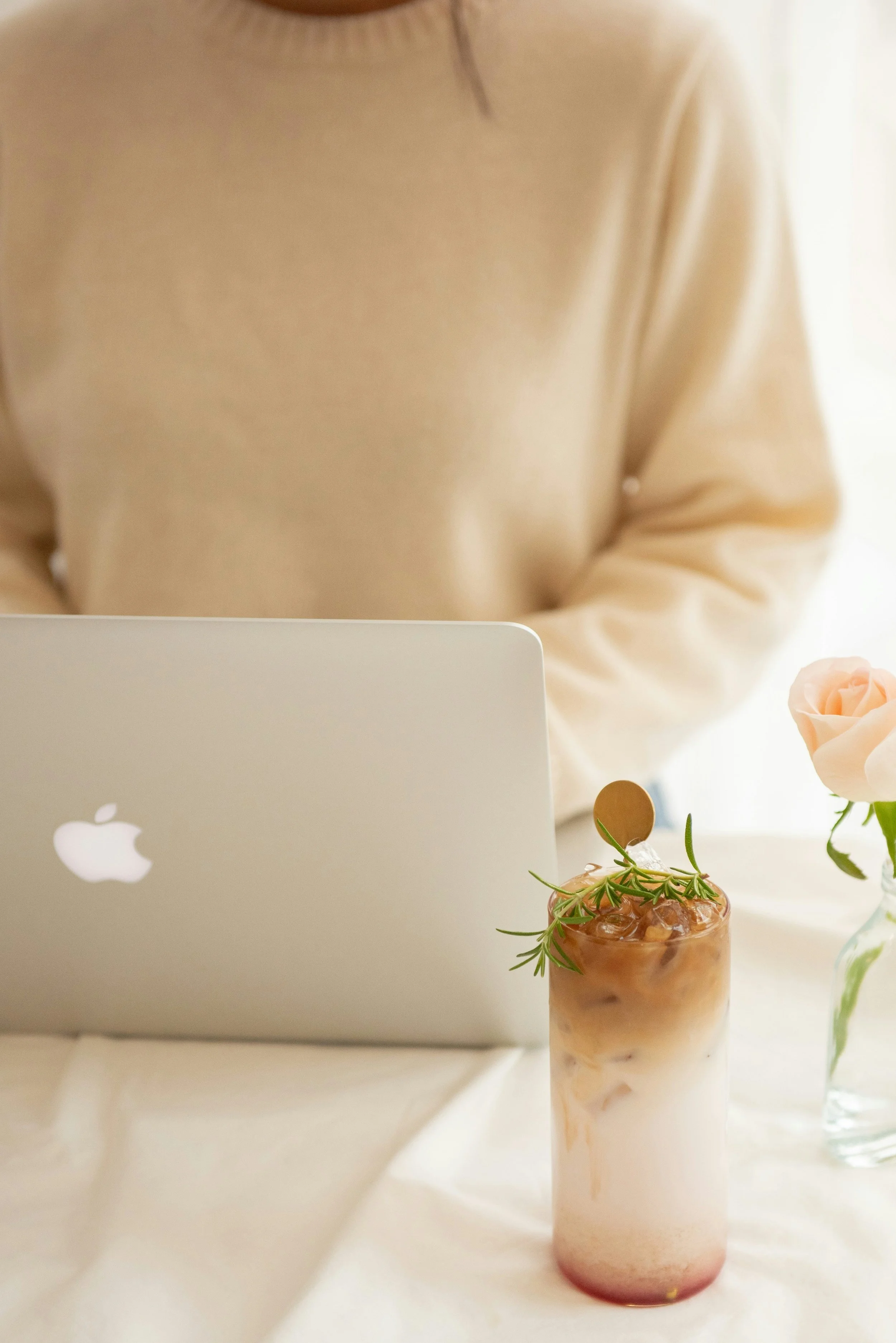 A person sitting at a table with a silver MacBook laptop, a pink and white marbled drink garnished with herbs, and a vase with a pink rose.