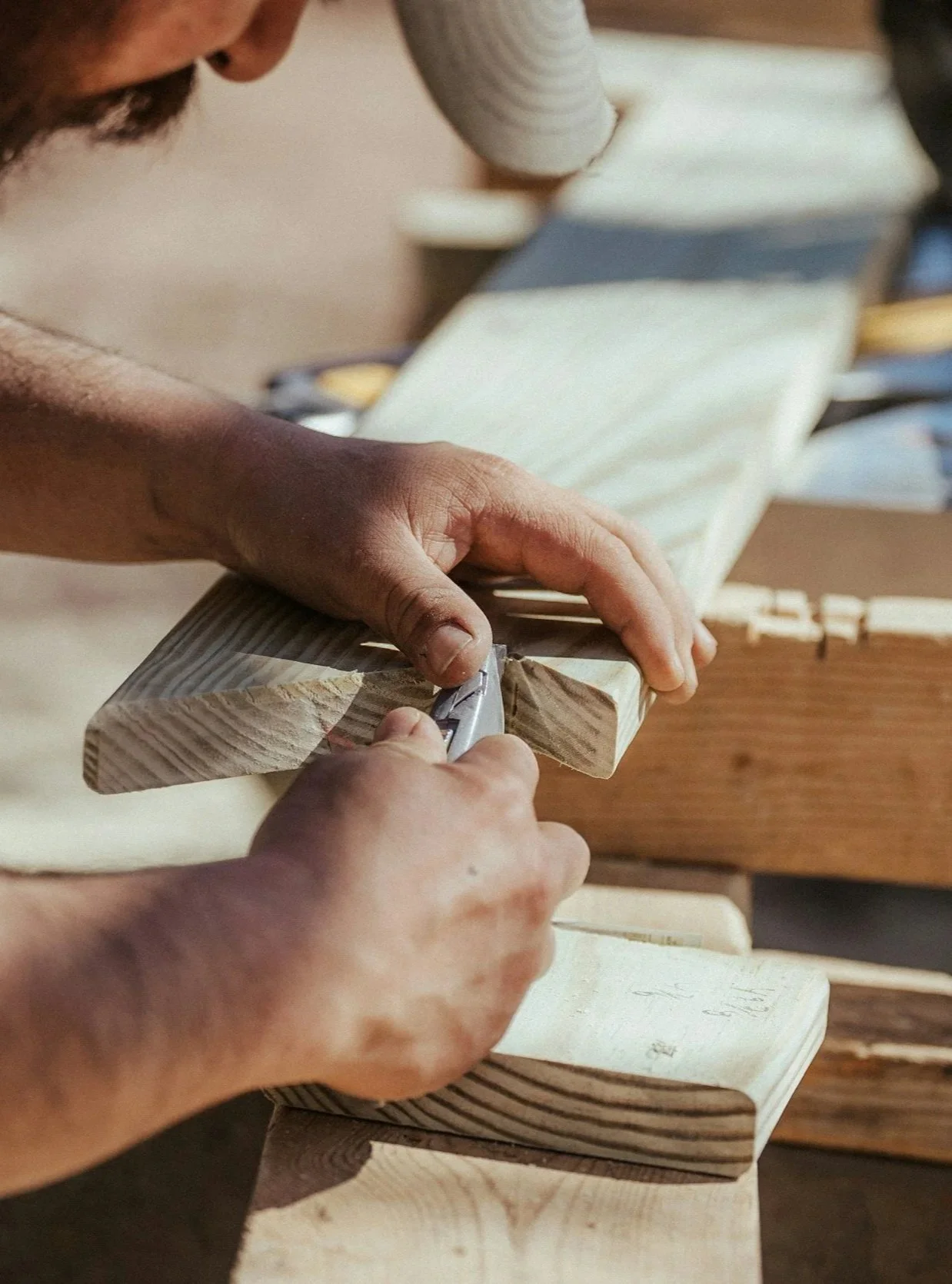 Close-up of a person using a utility knife to cut a piece of wood in a woodworking shop.