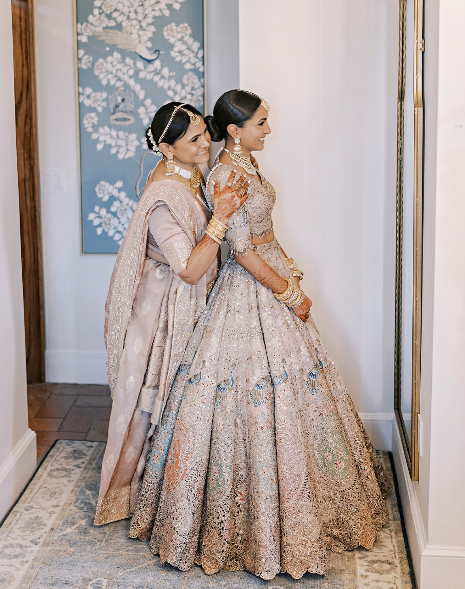 Two women dressed in traditional Indian bridal attire, standing side by side and smiling. The woman in the foreground wears a heavily embellished lehenga with intricate embroidery, and the woman behind her wears a similarly ornate saree or lehenga. T