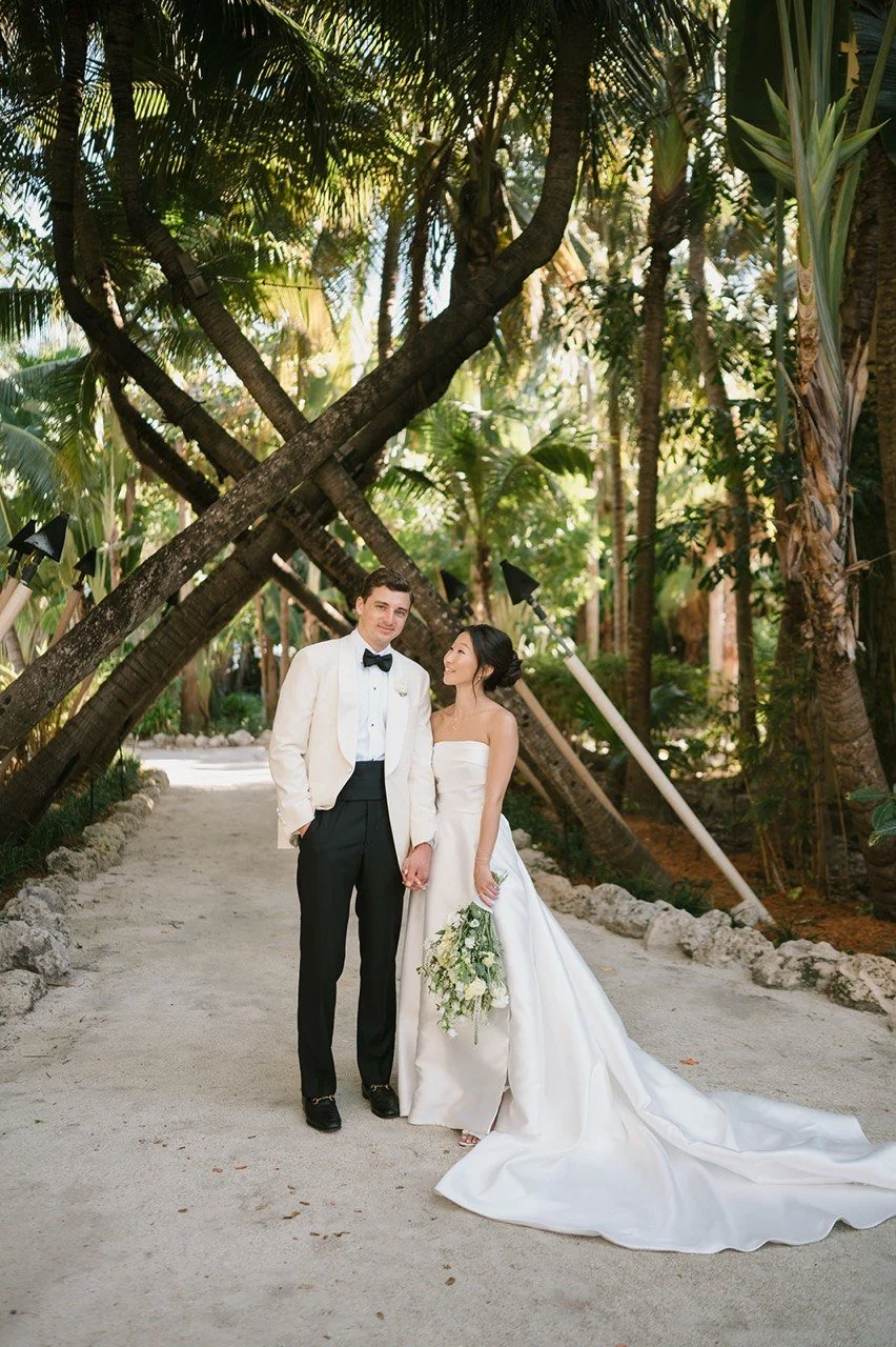 A bride and groom holding hands and standing on a sandy pathway in a lush tropical garden with large palm trees. The groom is wearing a black tuxedo with a white jacket and black bow tie, while the bride is wearing a strapless white wedding gown and 