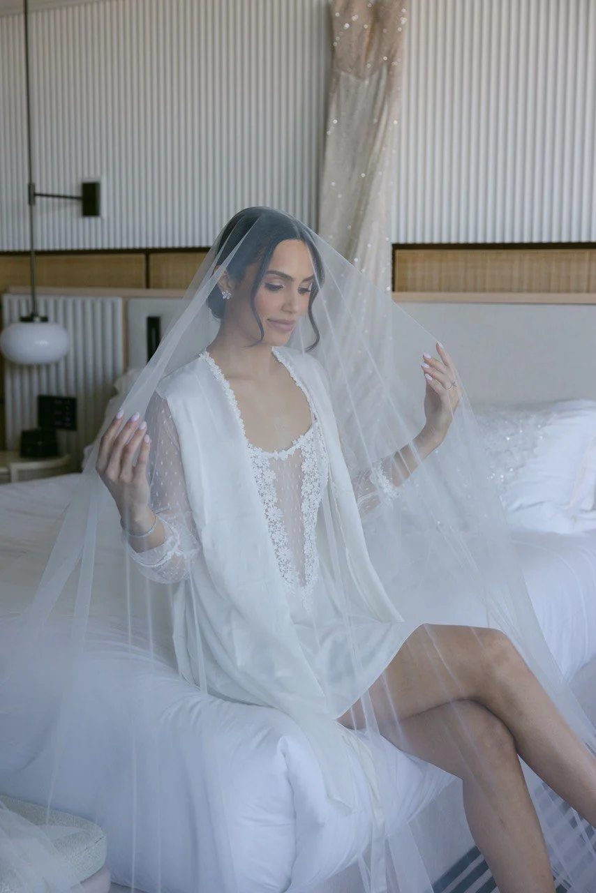 A bride sitting on a bed, adjusting her veil, dressed in white lace wedding attire with her eyes closed in a modern, minimalistic room.