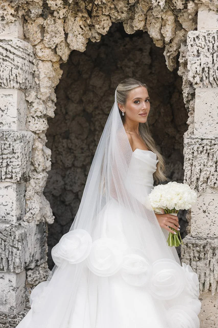 A bride in a white wedding gown with a veil, holding a bouquet of white flowers, standing in front of a rustic stone archway.