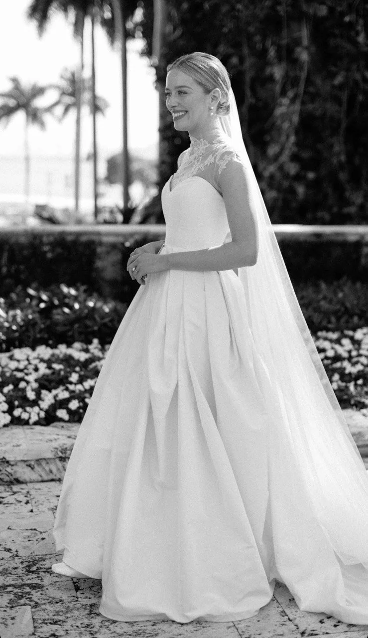 Black and white photo of a bride in a wedding dress standing outdoors, smiling, with palm trees and flowers in the background.