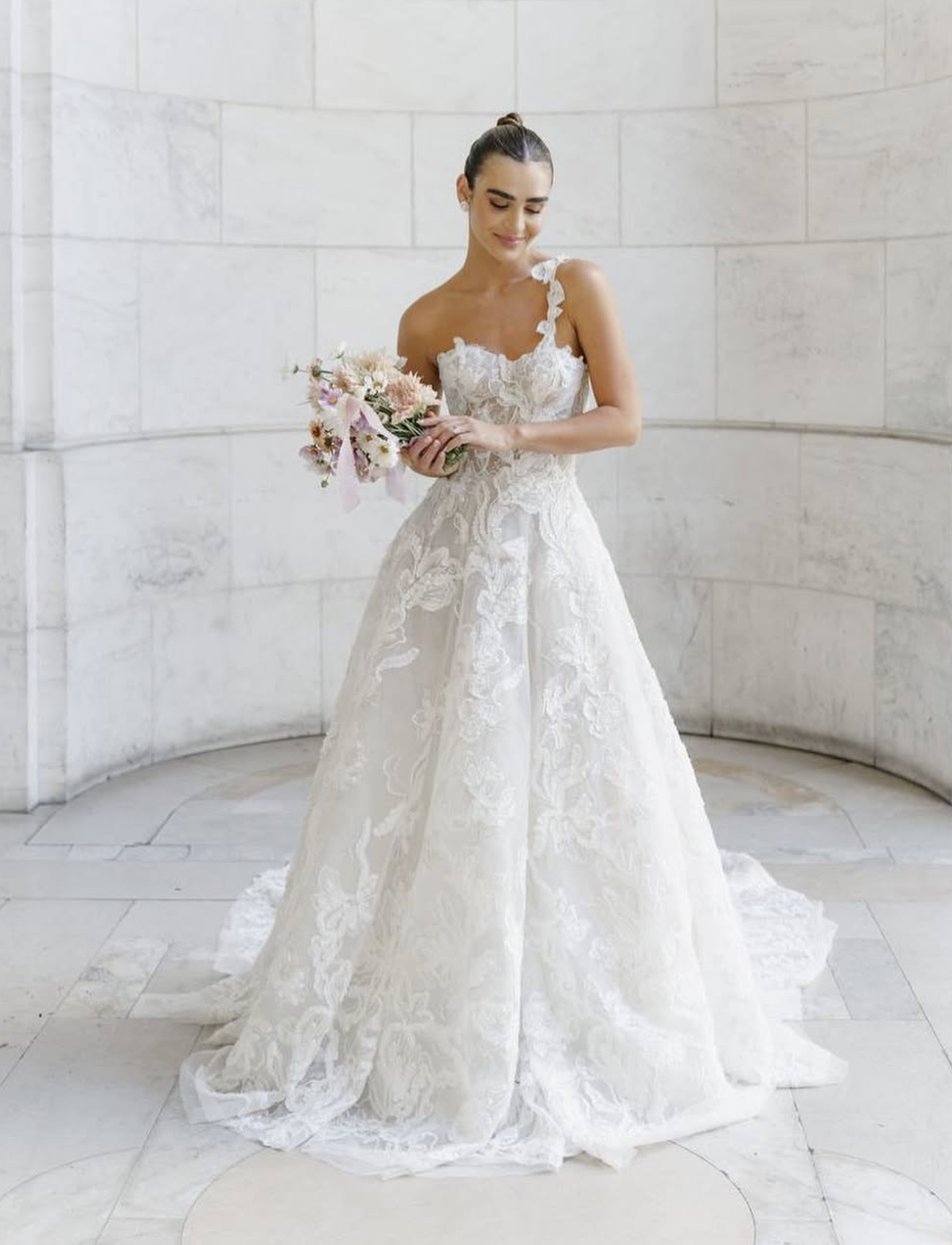 A bride standing indoors in front of a light-colored tiled wall, wearing a white lace wedding gown with a fitted bodice, holding a bouquet of flowers, and smiling gently.