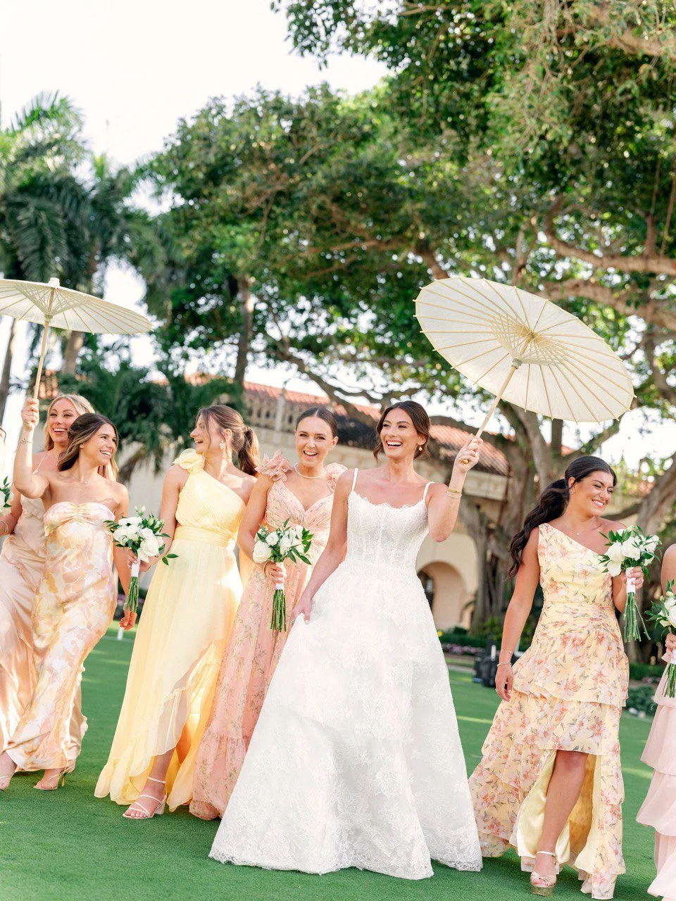 A bride in a white wedding dress walking outdoors with bridesmaids dressed in pastel-colored dresses, holding white bouquets and sunny parasols, under trees on a green lawn.