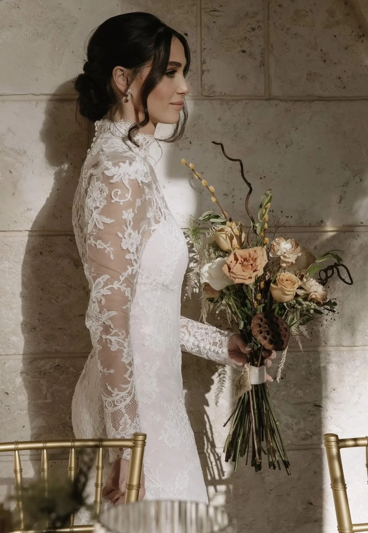 A woman in a white lace wedding gown holding a bouquet of flowers against a stone wall.