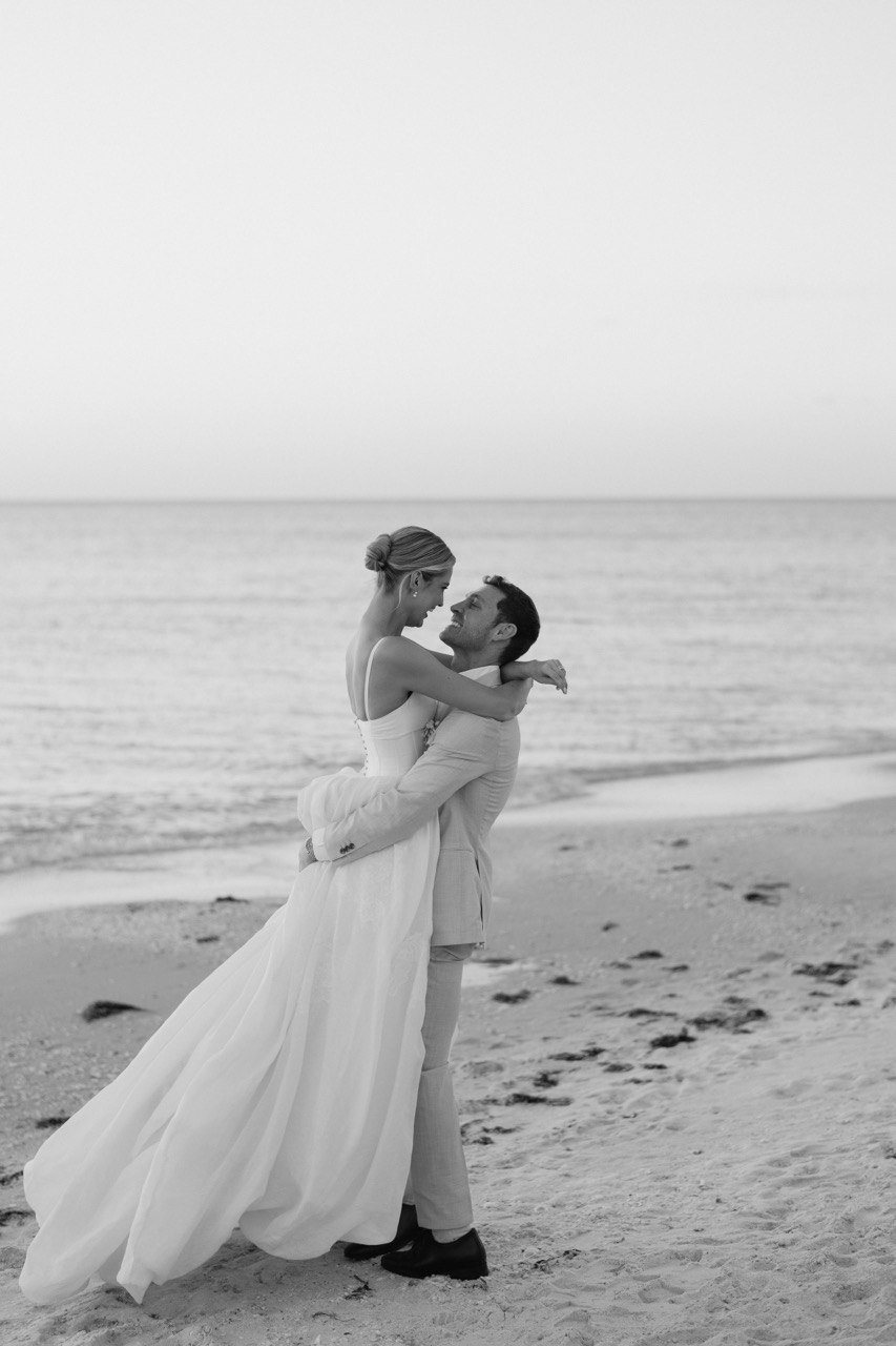A black and white photo of a couple on a beach, with the man lifting the woman who is wearing a wedding dress, and they are looking into each other's eyes.