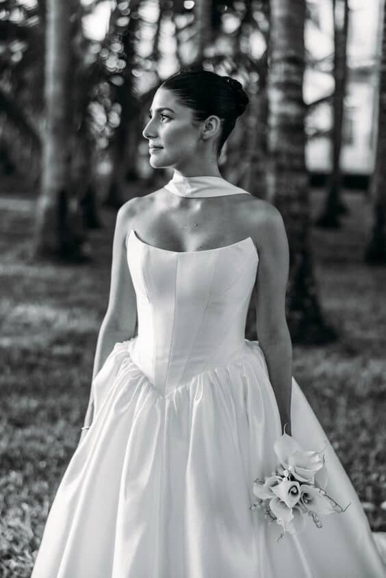 A woman in a strapless wedding dress holding a bouquet of roses, standing outdoors with trees in the background.