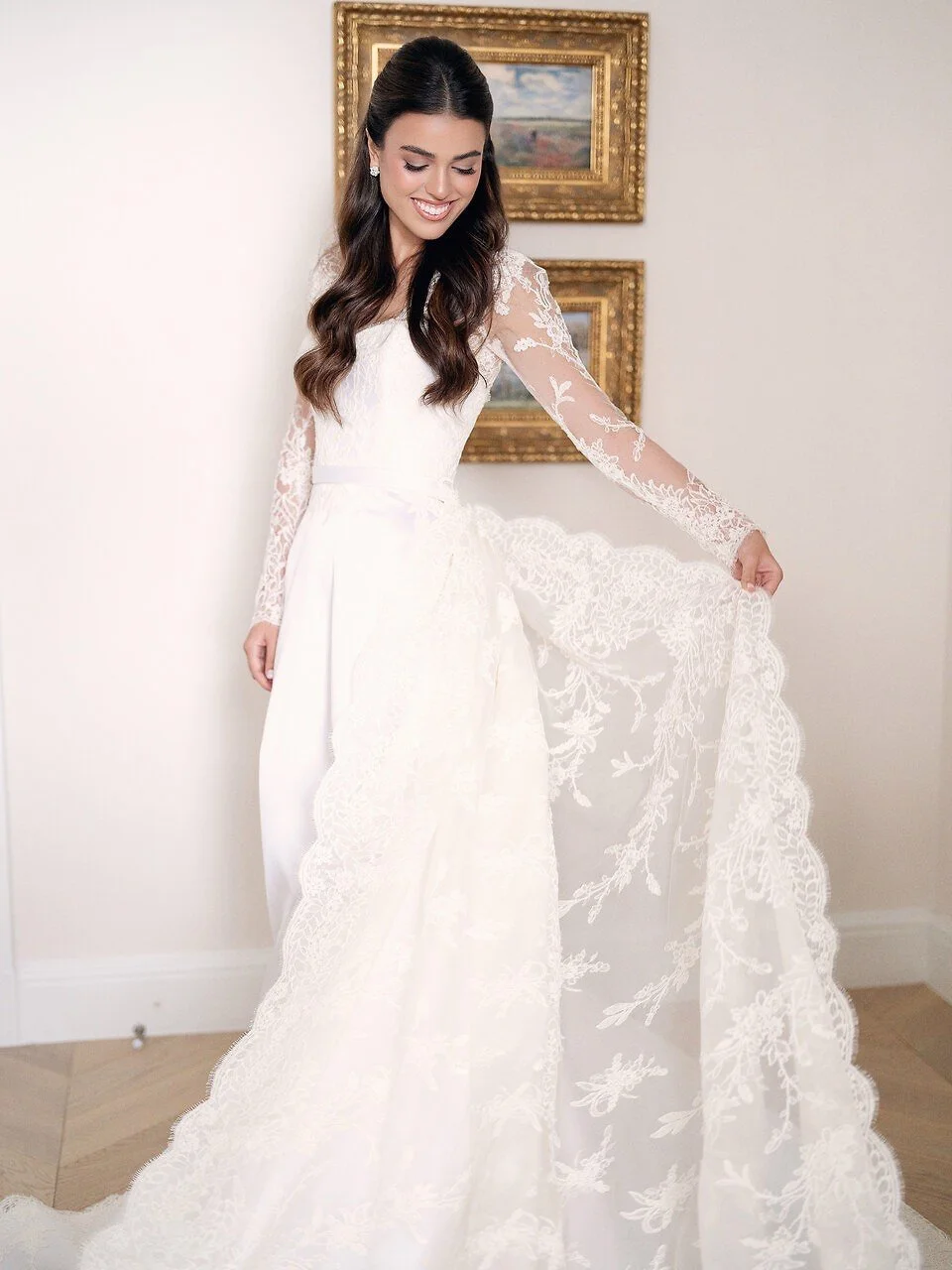 A woman in a white wedding dress with lace sleeves, smiling and holding out the train of her dress, standing in front of a white wall with framed landscape paintings.