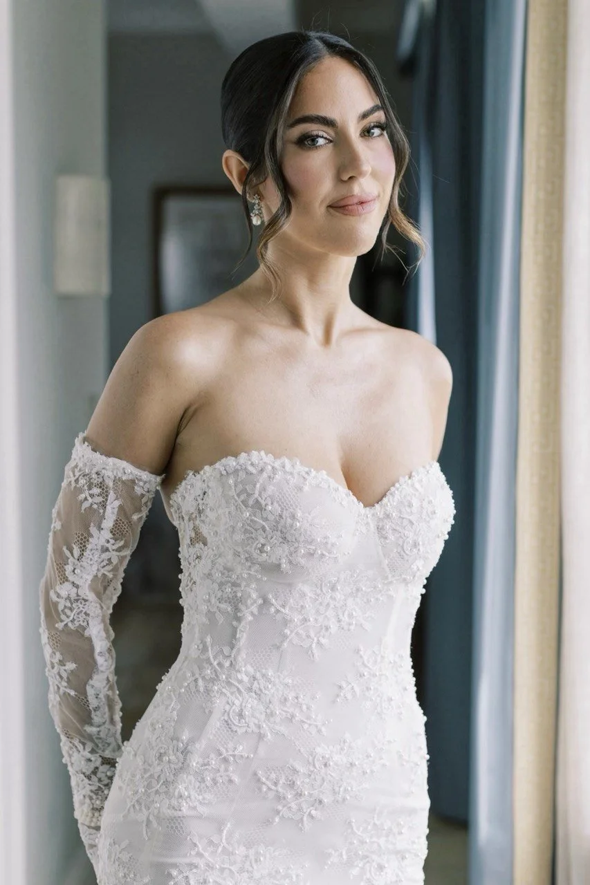 A woman in a white wedding dress with lace sleeves and floral embroidery, standing near a window.