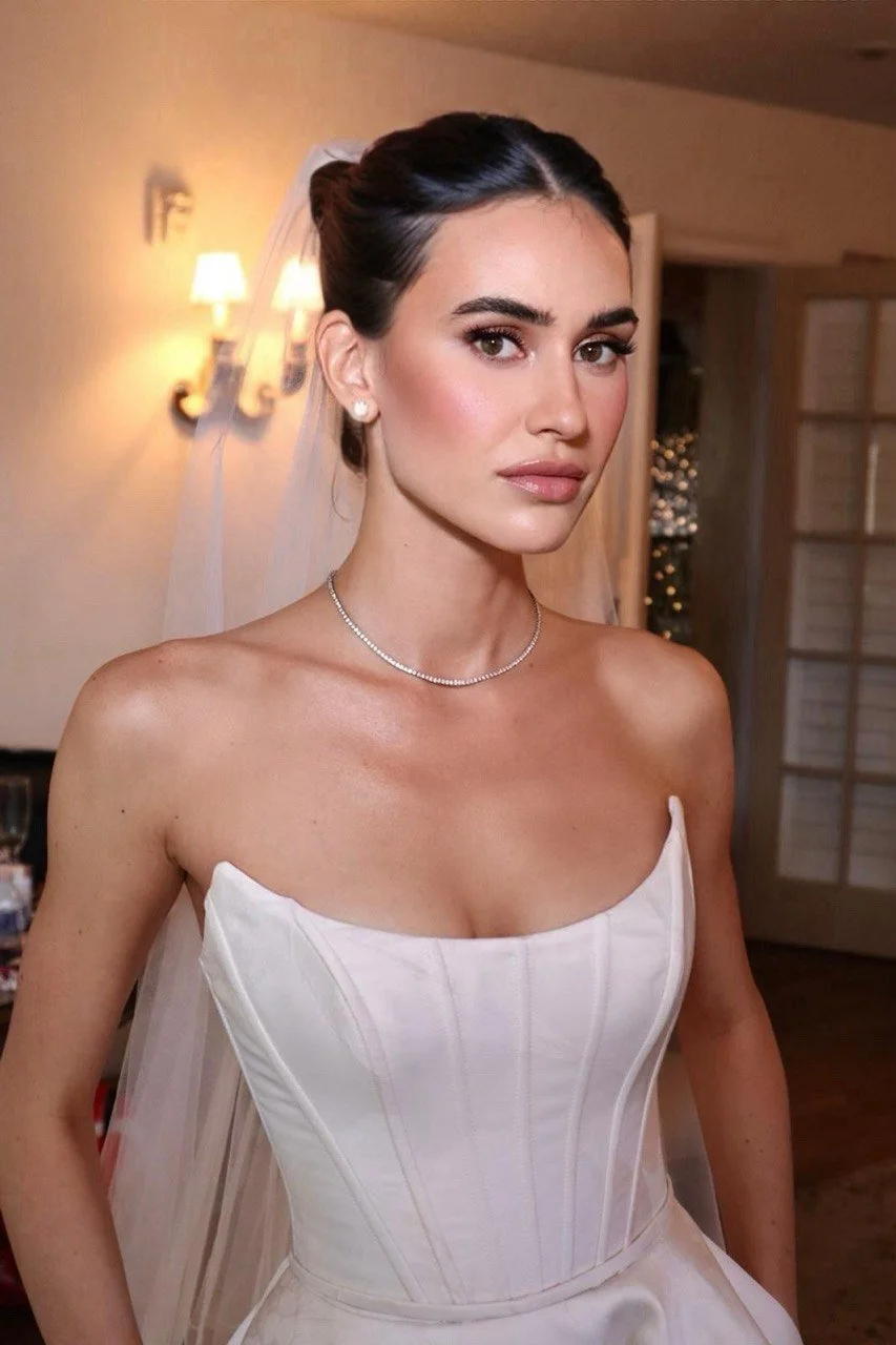 A young woman with dark hair styled in an elegant updo, wearing a white strapless wedding gown and matching veil, standing indoors with warm lighting and a background featuring a wall sconce and a room with wooden furniture.