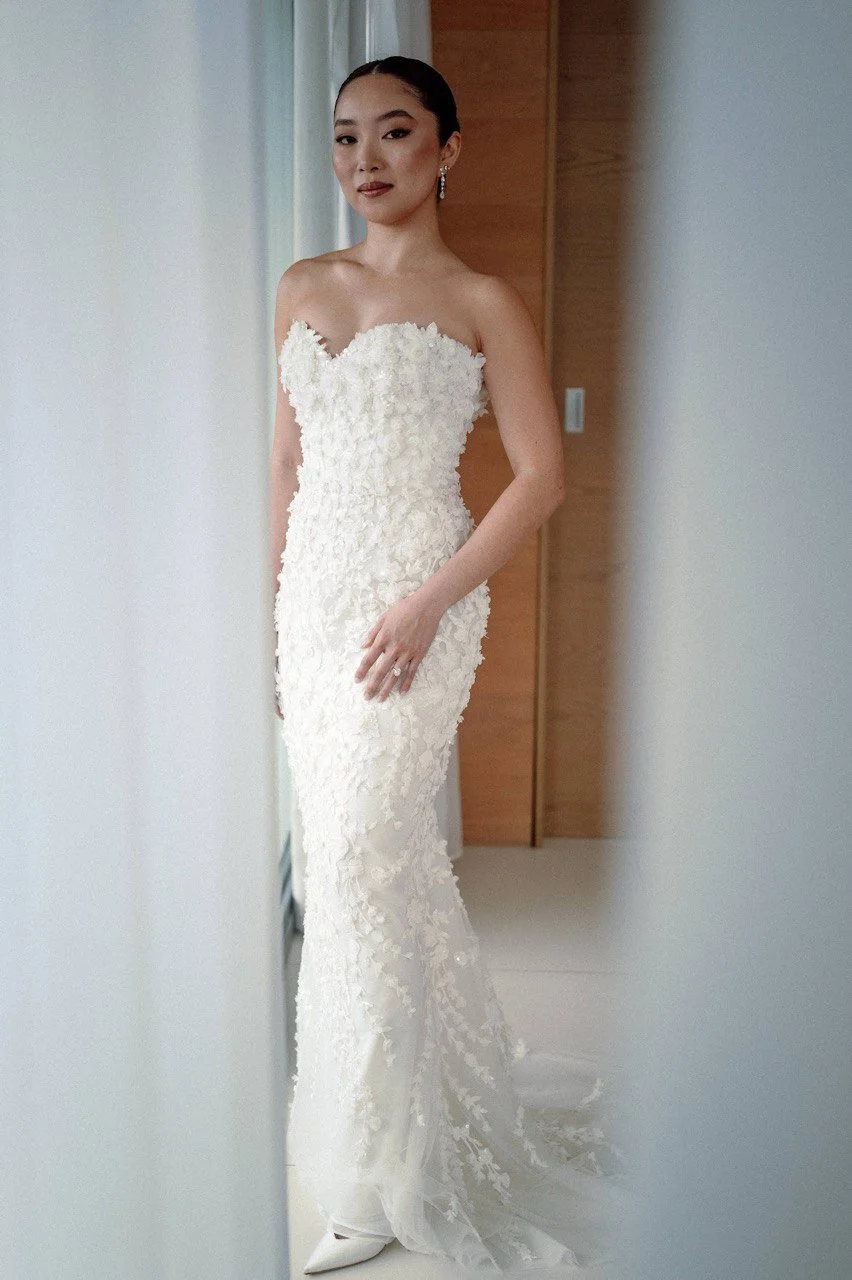 Woman in a strapless white wedding gown with floral lace details, standing in a room with wooden panel and white wall, looking at the camera.