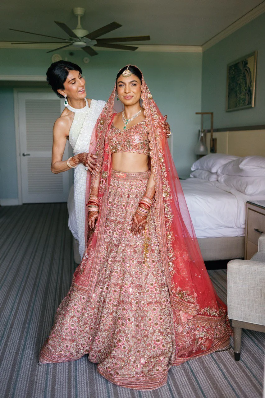 An Indian bride in a pink and gold embroidered lehenga and matching dupatta standing in a hotel room, with another woman helping her adjust the attire.