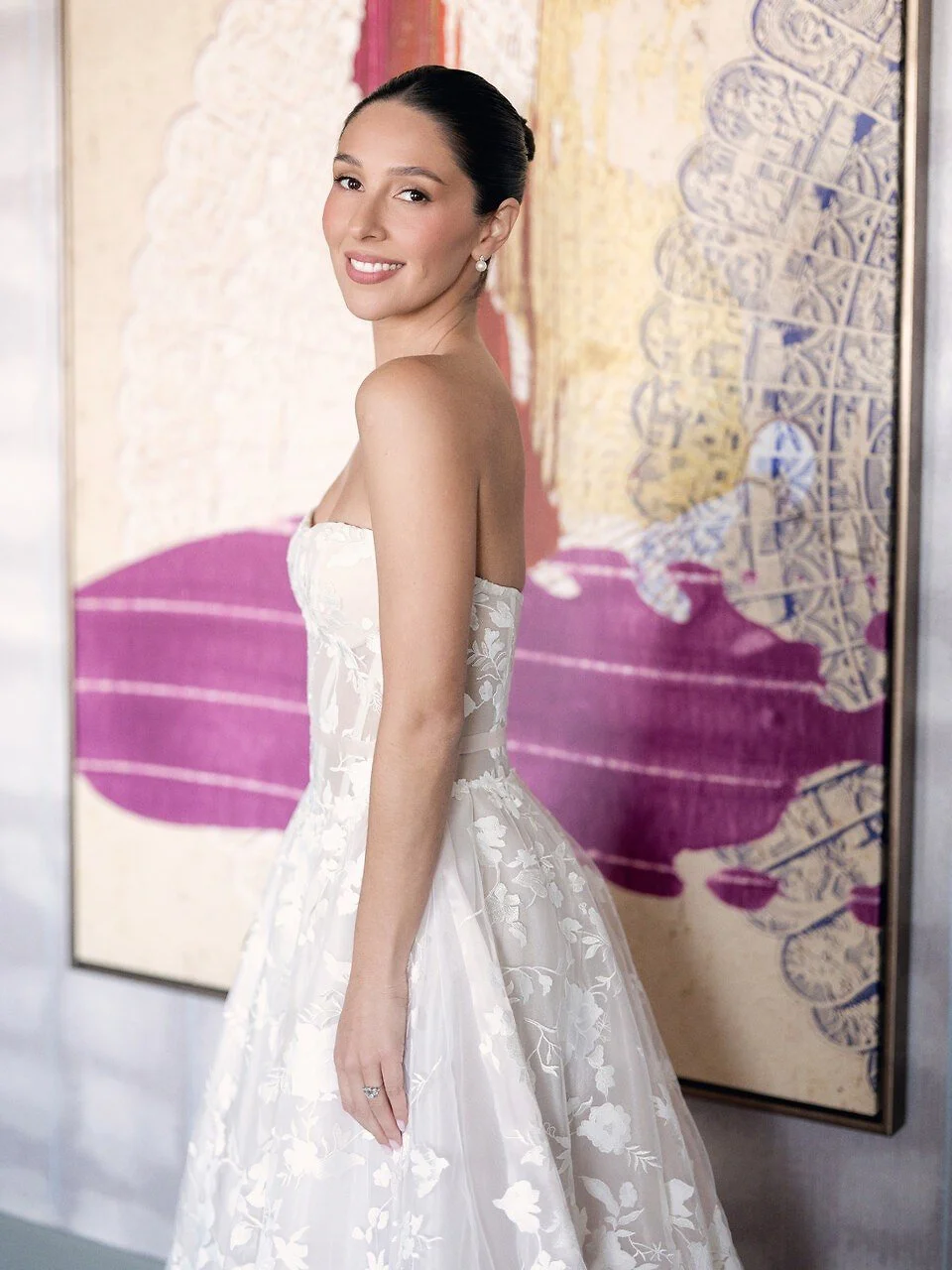 A woman in a white strapless wedding gown with floral embroidery, smiling and standing indoors against a colorful abstract background.