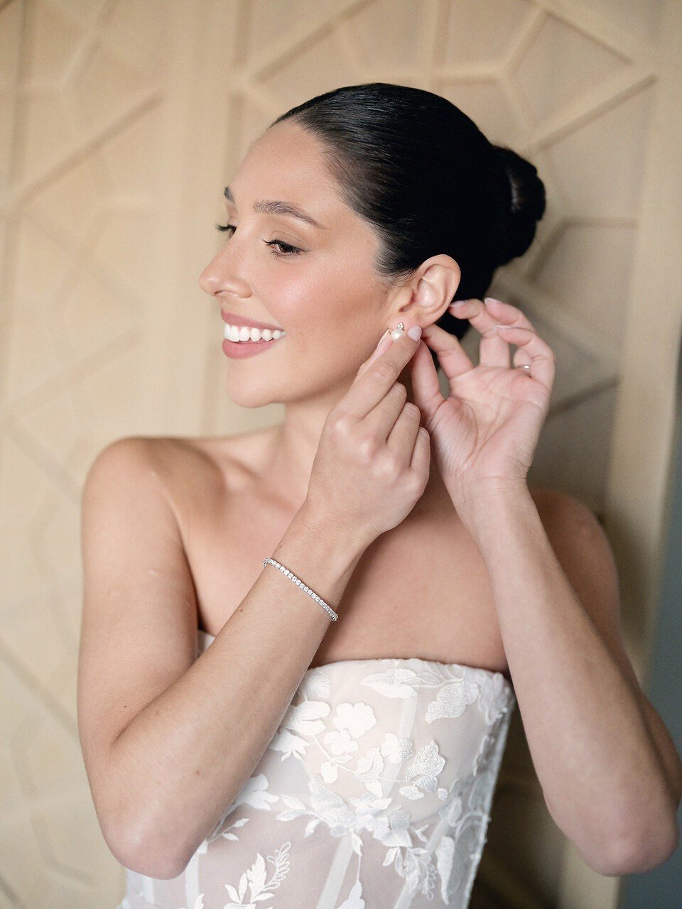 A smiling woman dressed in a strapless floral wedding gown, putting on earrings.