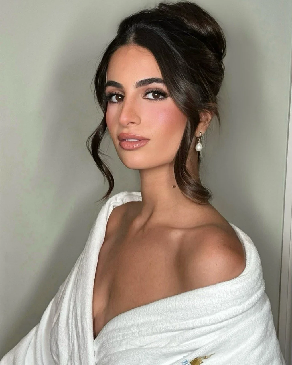 A woman with dark hair styled in loose curls and an elegant updo, wearing pearl drop earrings and a white off-shoulder top, posing against a plain wall.