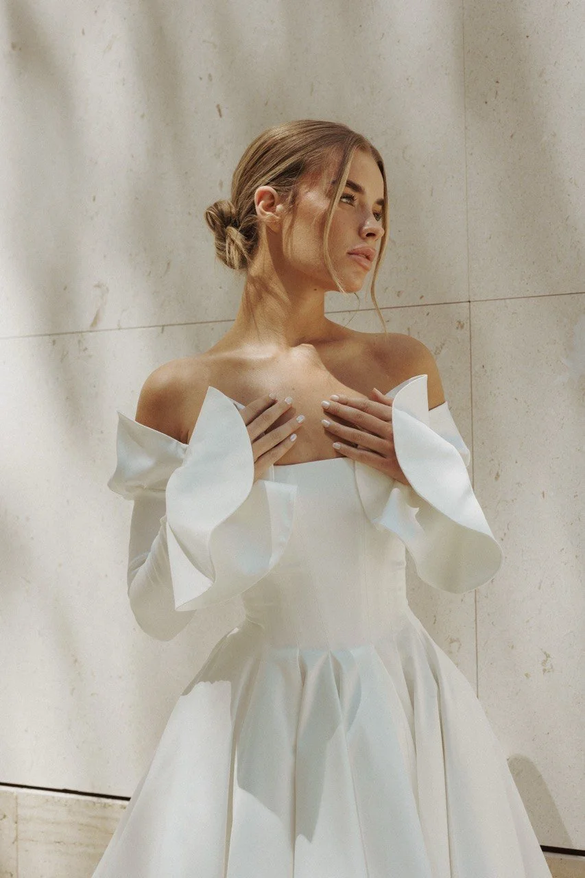 A young woman in a white, off-the-shoulder dress with ruffled sleeves, holding her chest with both hands, standing against a light-colored wall, with a thoughtful expression.