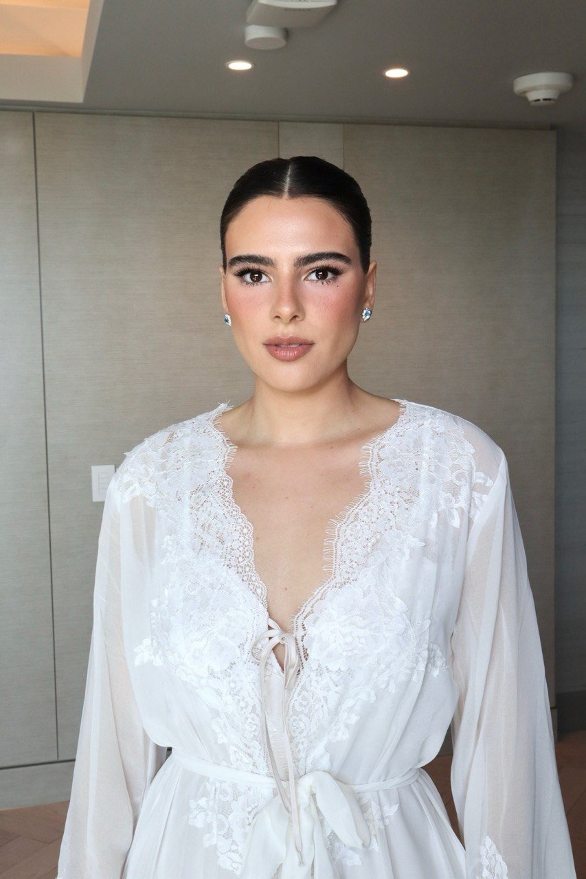 A woman with dark hair pulled back, wearing a white lace and satin robe with a front tie, standing indoors with neutral-colored walls and ceiling lights.