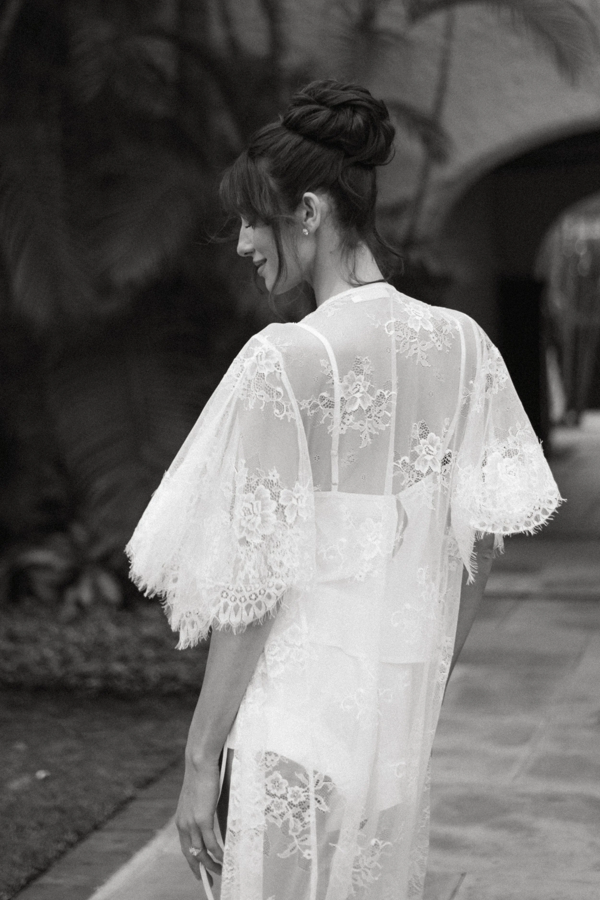 Black and white image of a woman wearing a lace robe, side profile, with her hair styled in an elegant updo, standing outdoors near a building and trees.