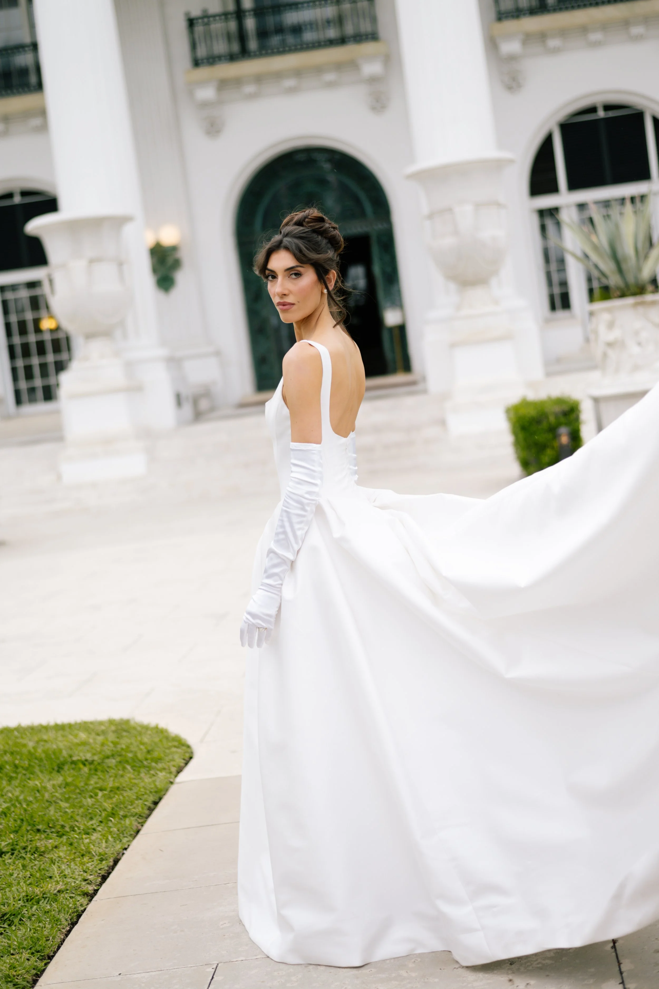 A woman in a white wedding dress with long gloves stands outside a white building with columns and large arched windows.