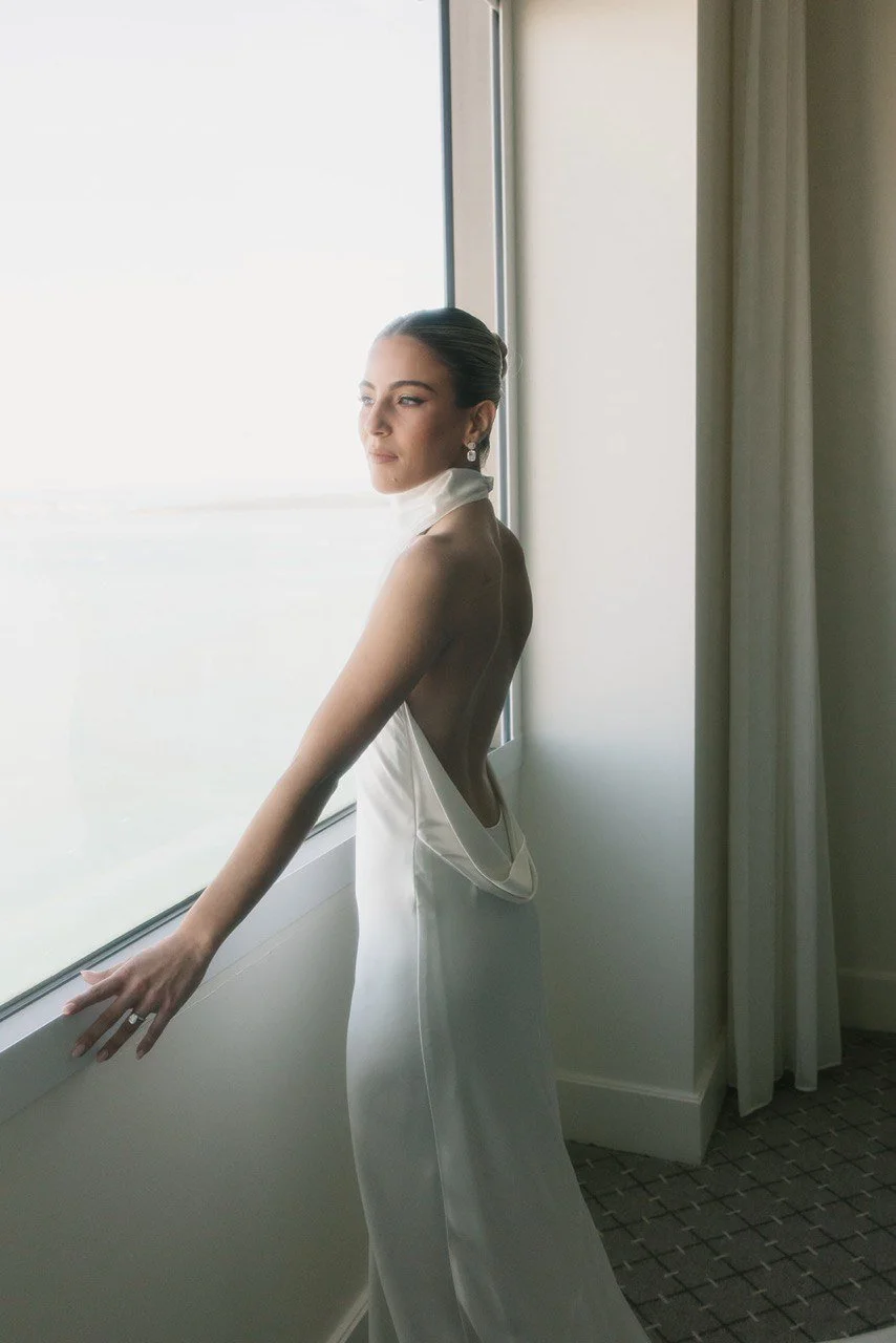 A woman in a white dress looking out of a window, standing in a room with curtains and a patterned carpet.