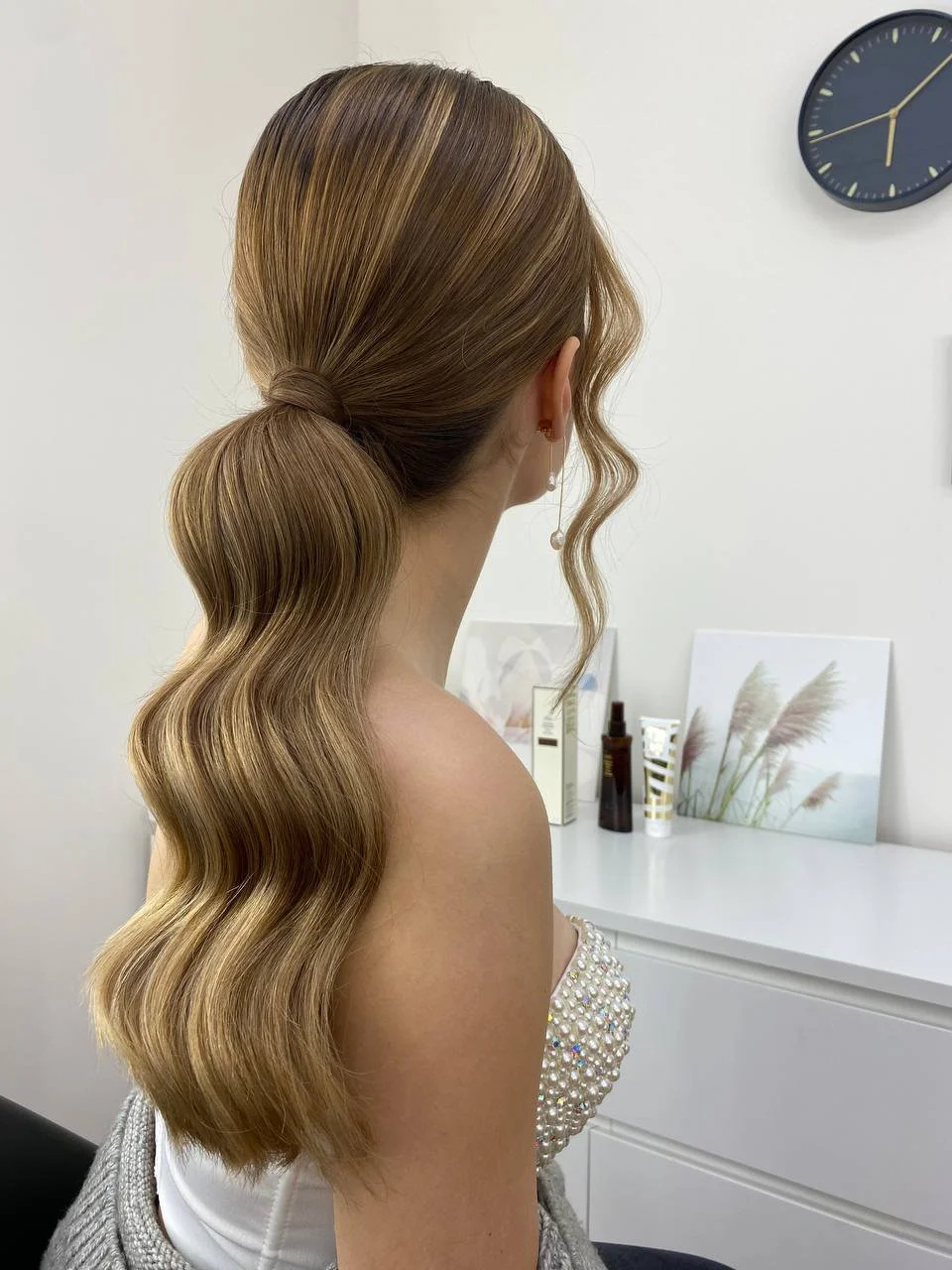 Woman with long, wavy blonde hair tied in a low ponytail, wearing a pearl-embellished top and a dangling pearl earring, inside a room with a white wall, a clock, and decorative items on a white dresser.