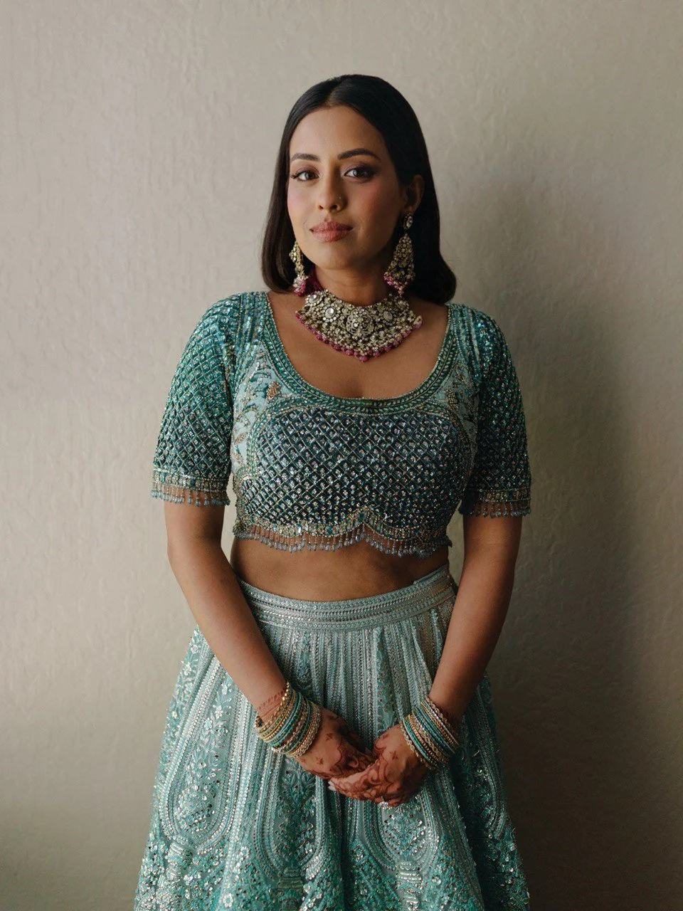 A woman wearing traditional Indian jewelry and a teal embroidery blouse and skirt, standing against a plain wall.