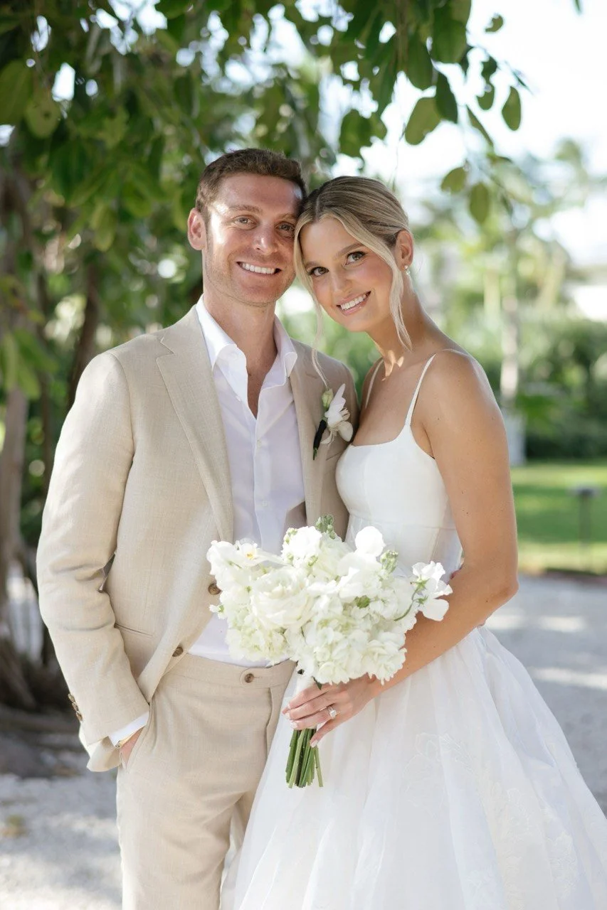 A newlywed couple dressed in wedding attire, standing outdoors beneath a tree, smiling and holding a bouquet of white flowers.