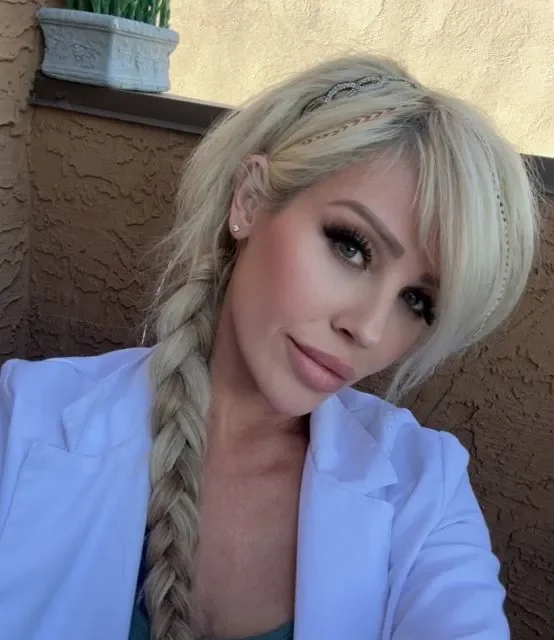 A woman with long blonde hair styled in a braid, wearing makeup and a white top, taking a selfie outdoors against a textured brown wall with a window flower box in the background.