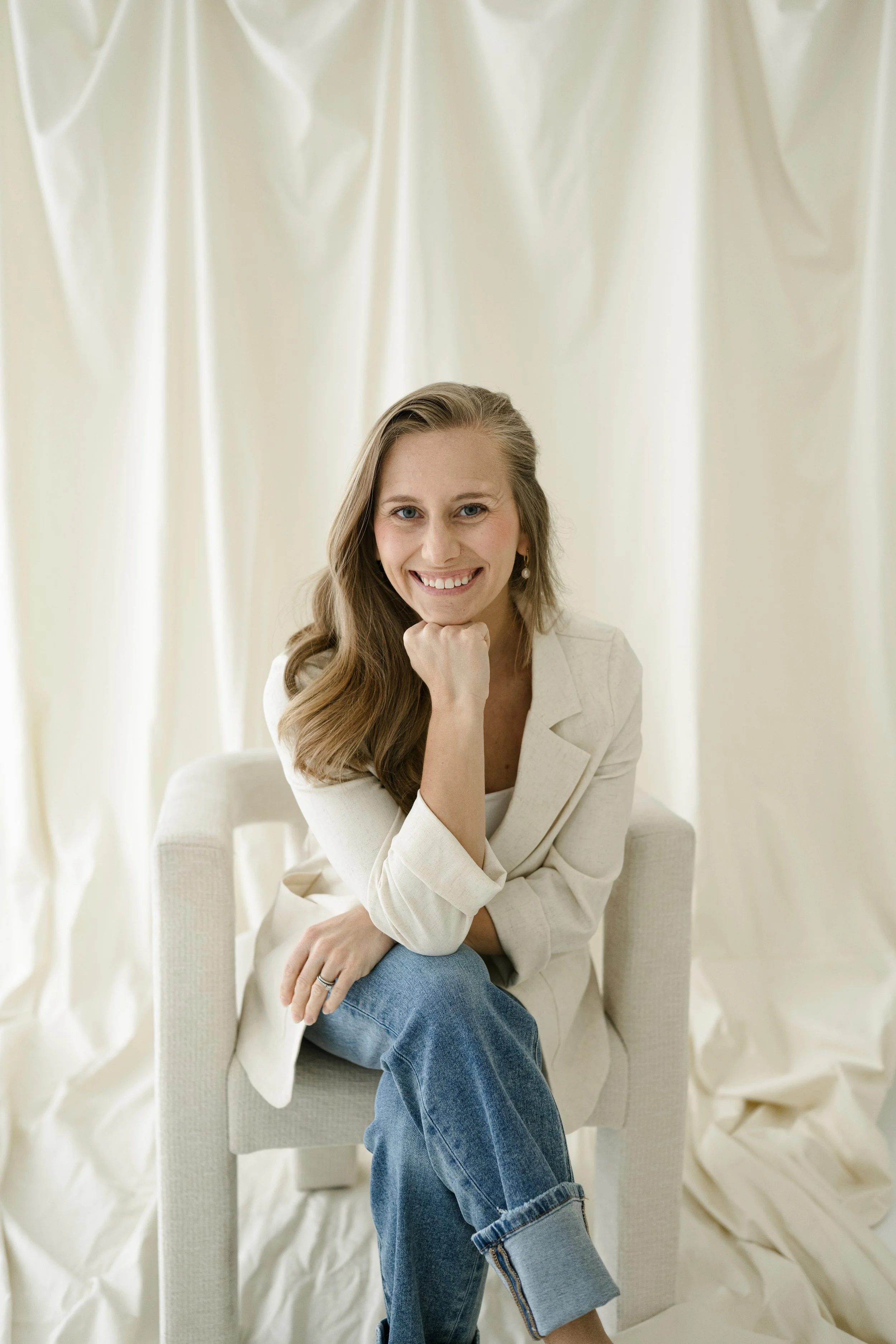 A young woman with long wavy hair, wearing a white blazer and blue jeans, sitting on a cream-colored armchair in front of cream-colored curtains, smiling and resting her chin on her hand.