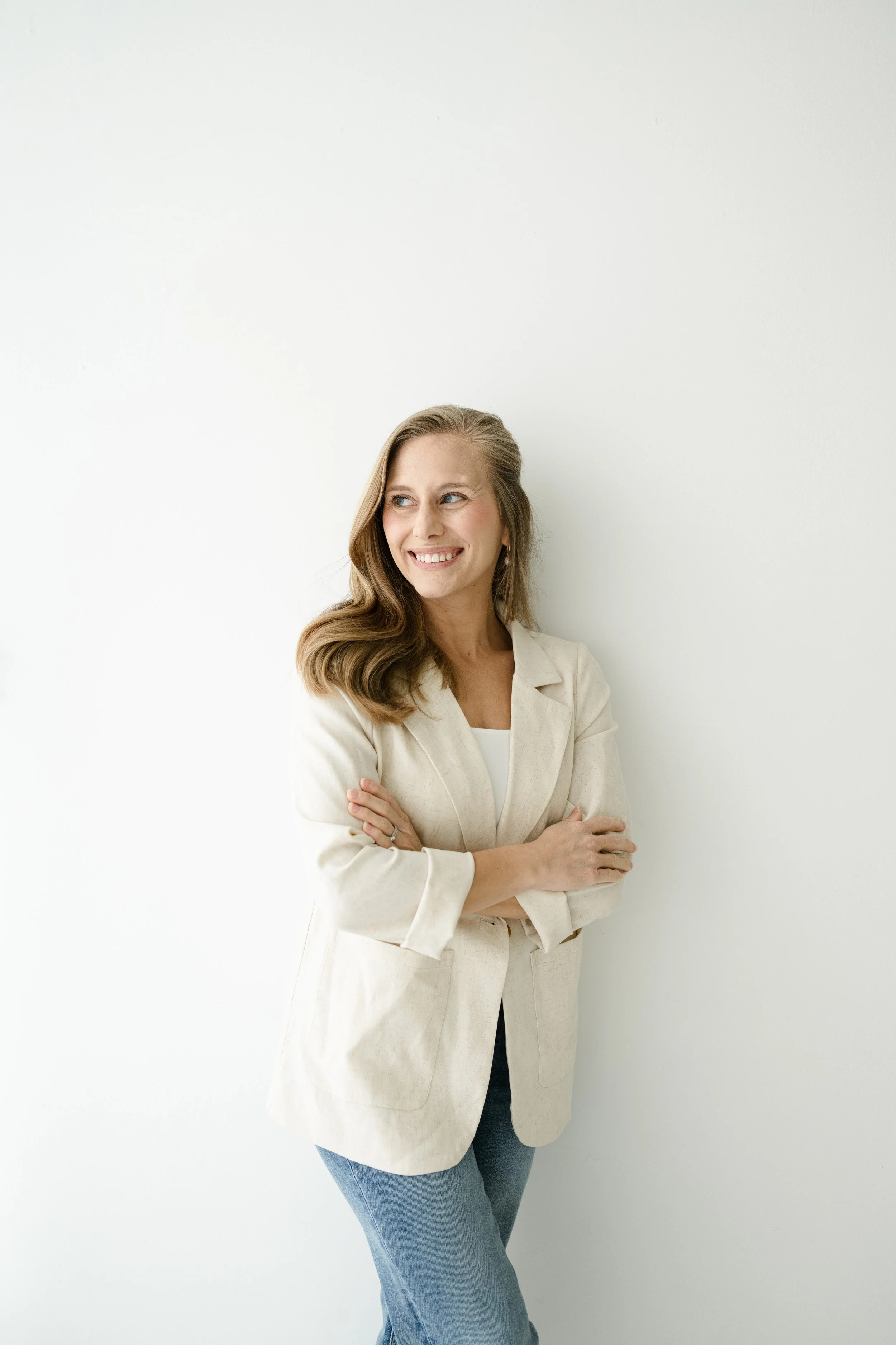 A woman with long wavy hair smiling and looking to her right, wearing a light-colored blazer and blue jeans, standing against a plain white wall.