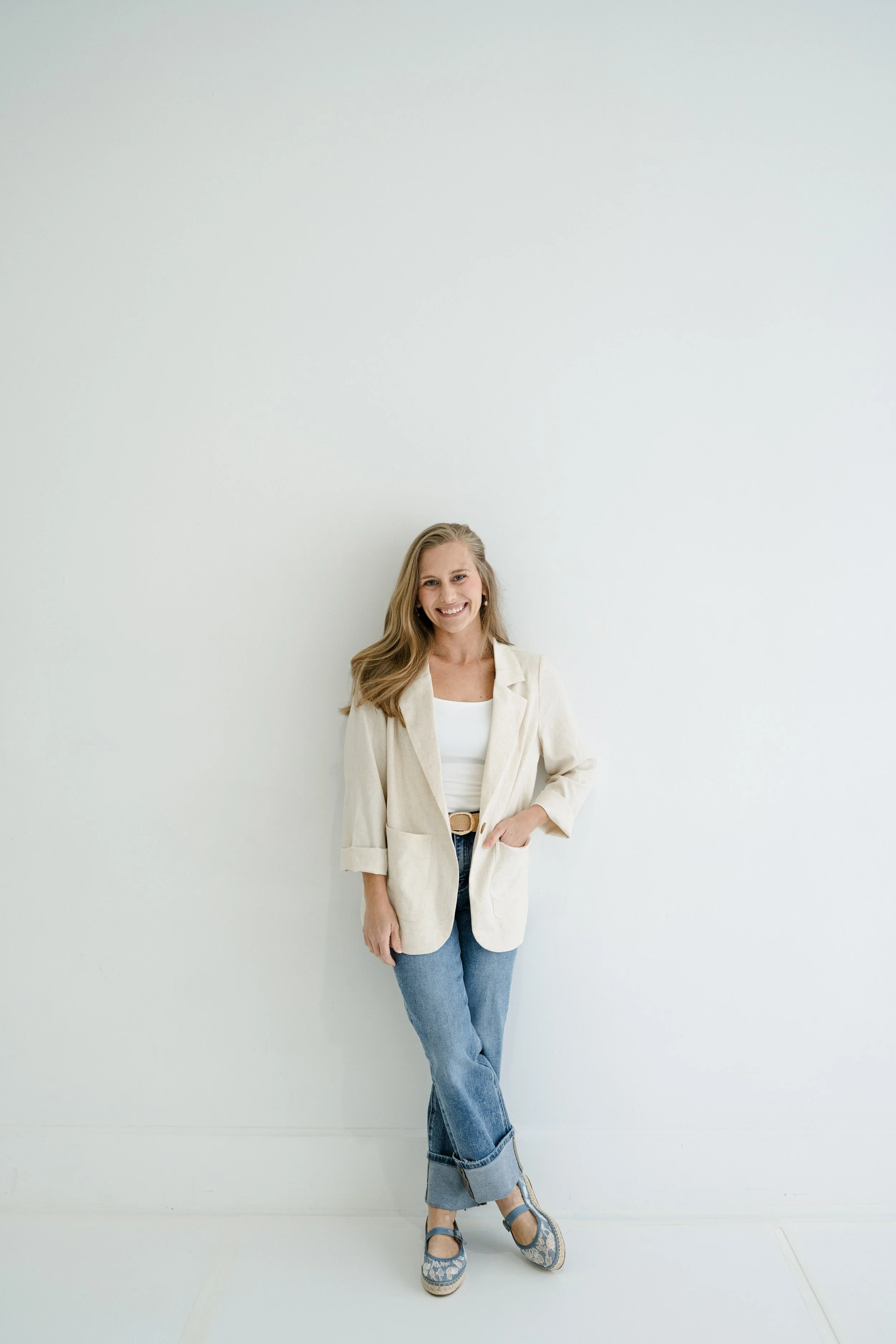 A woman standing against a plain white wall, smiling, wearing a light-colored blazer, white top, blue jeans, and blue patterned shoes.