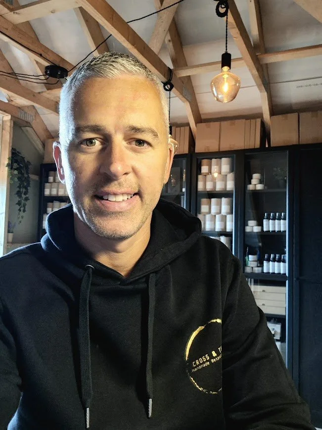 A man with short light hair smiling, wearing a black hoodie with a logo, standing inside a wooden ceiling space with glass cabinets filled with white containers and bottles.