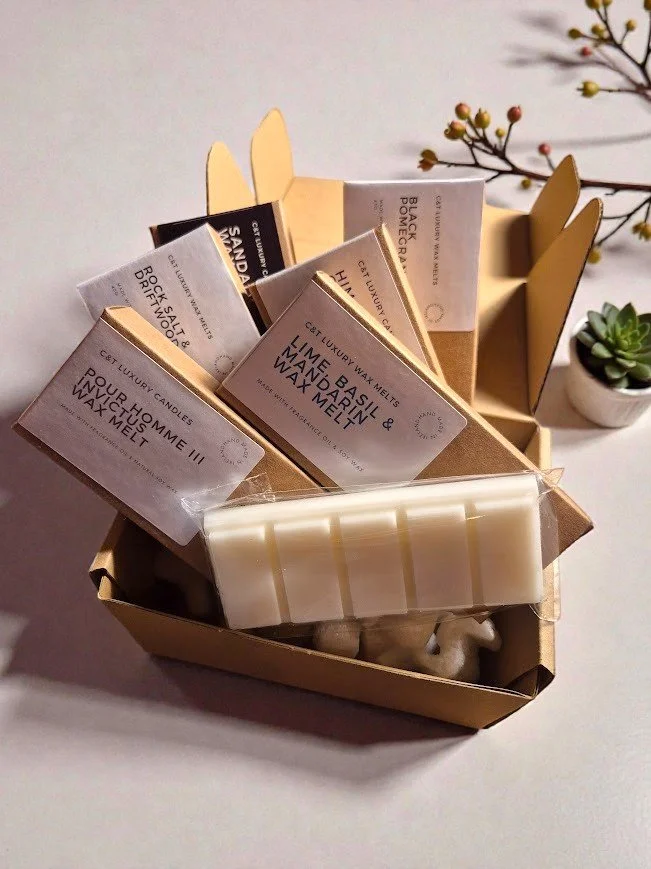 A cardboard storage box with a butterfly-shaped lid contains various scented wax melts and candles, with labels indicating flavors, along with a white wax block. There is a small potted succulent and a branch with tiny berries in the background.