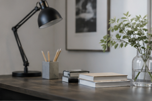 Office desk with black adjustable desk lamp, stack of books, gray pencil holder with pencils, and a glass vase with green plant, against gray wall with framed black and white photo.