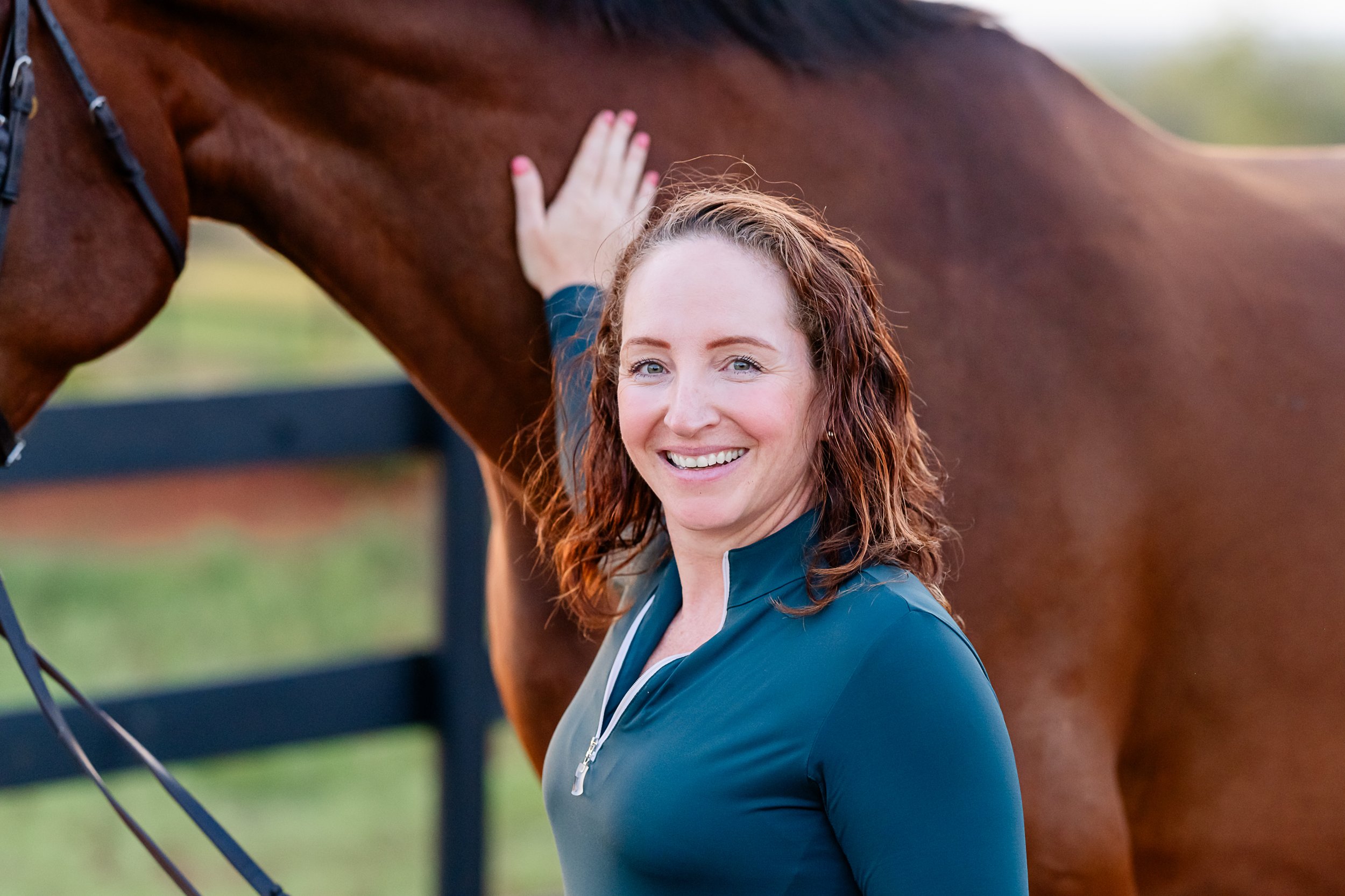 A smiling woman with curly reddish-brown hair, wearing a dark teal zip-up jacket, standing next to a chestnut-colored horse and petting its neck.