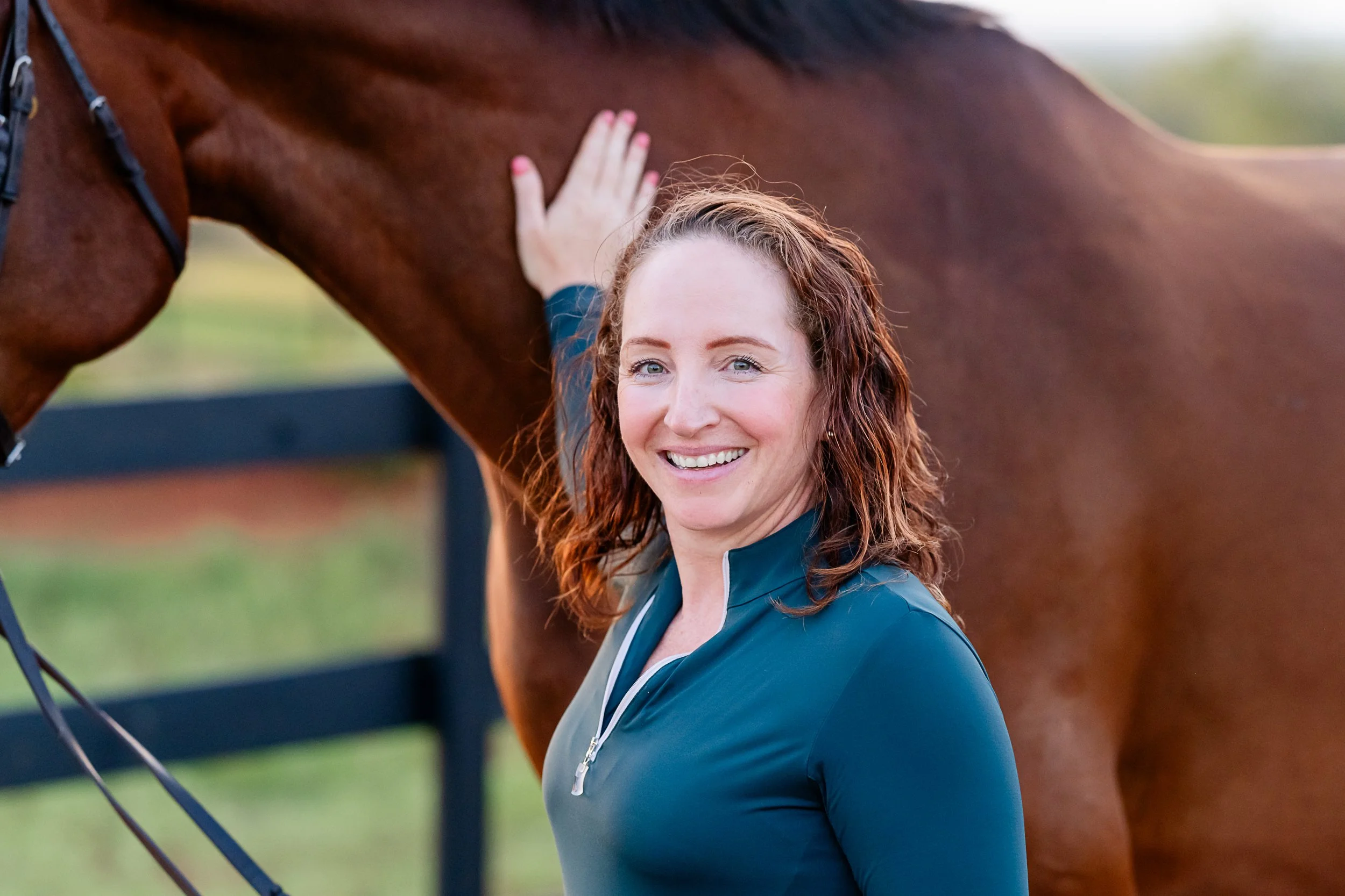 A smiling woman with curly reddish-brown hair, wearing a dark teal zip-up jacket, standing next to a chestnut-colored horse and petting its neck.