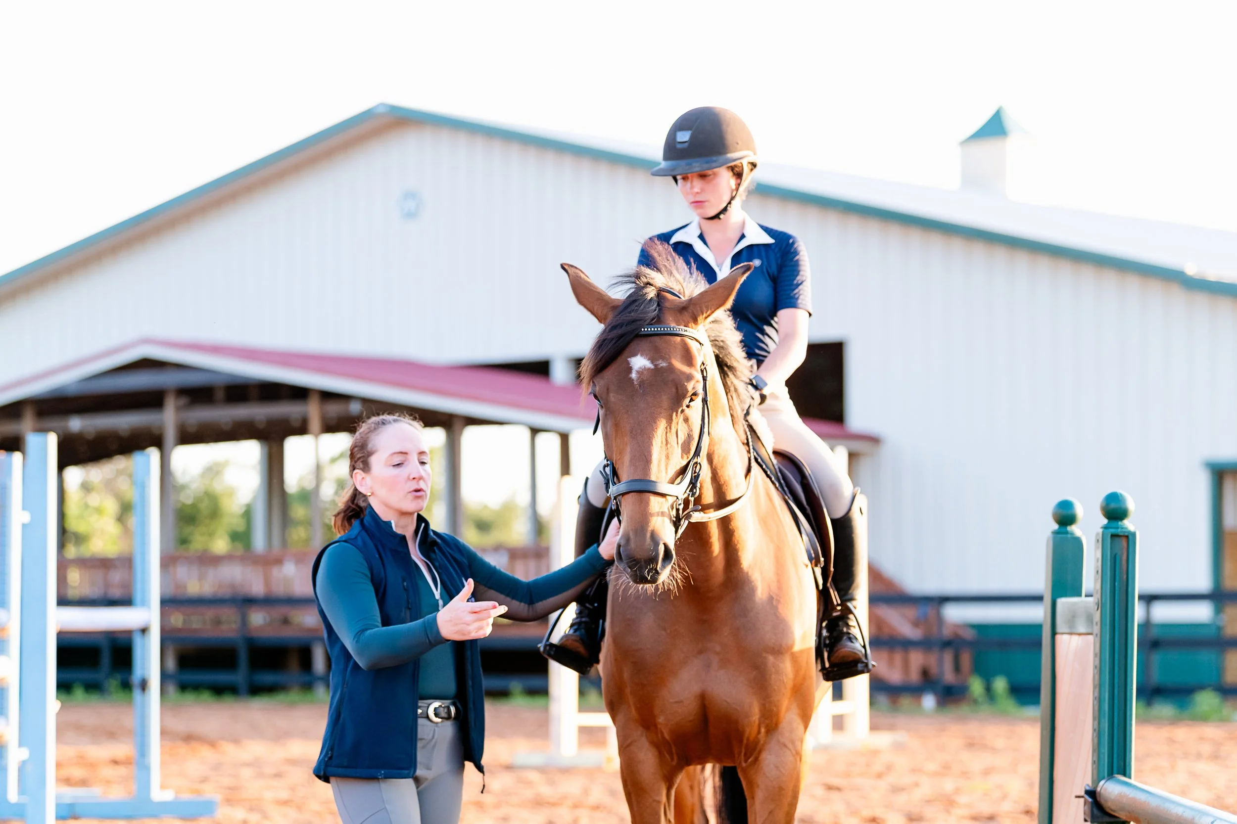 An adult riding student is sitting on a brown horse, and her trainer is standing next to them, in a riding lesson.
