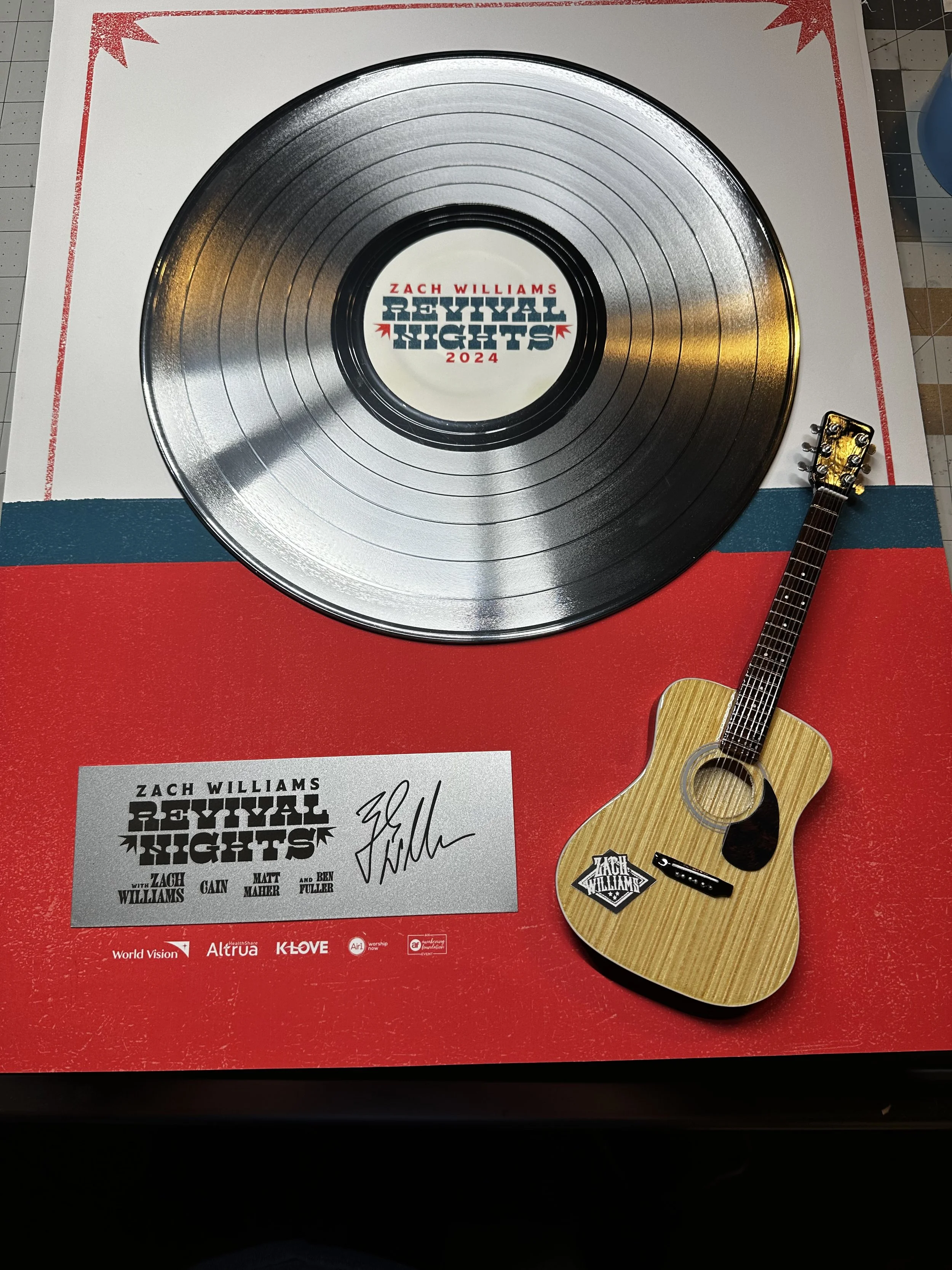 A vinyl record with the title 'Revivial Nights 2024' by Zach Williams in the center, placed on a red, white, and blue mat. There is a small acoustic guitar with a Zach Williams sticker, a signature, and several other signatures nearby.