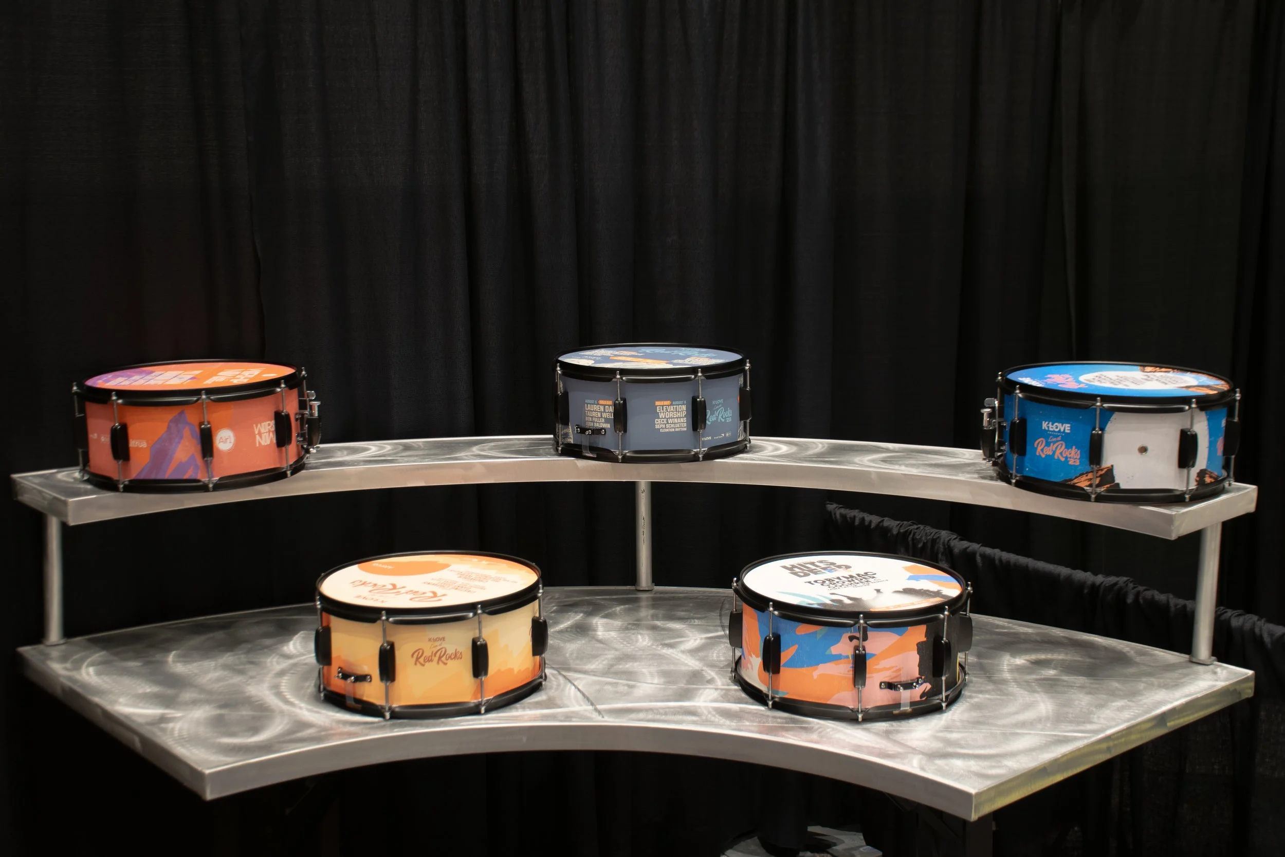Five snare drums with colorful promotional designs and text on top, placed on a two-tier metal display stand with a black curtain background.