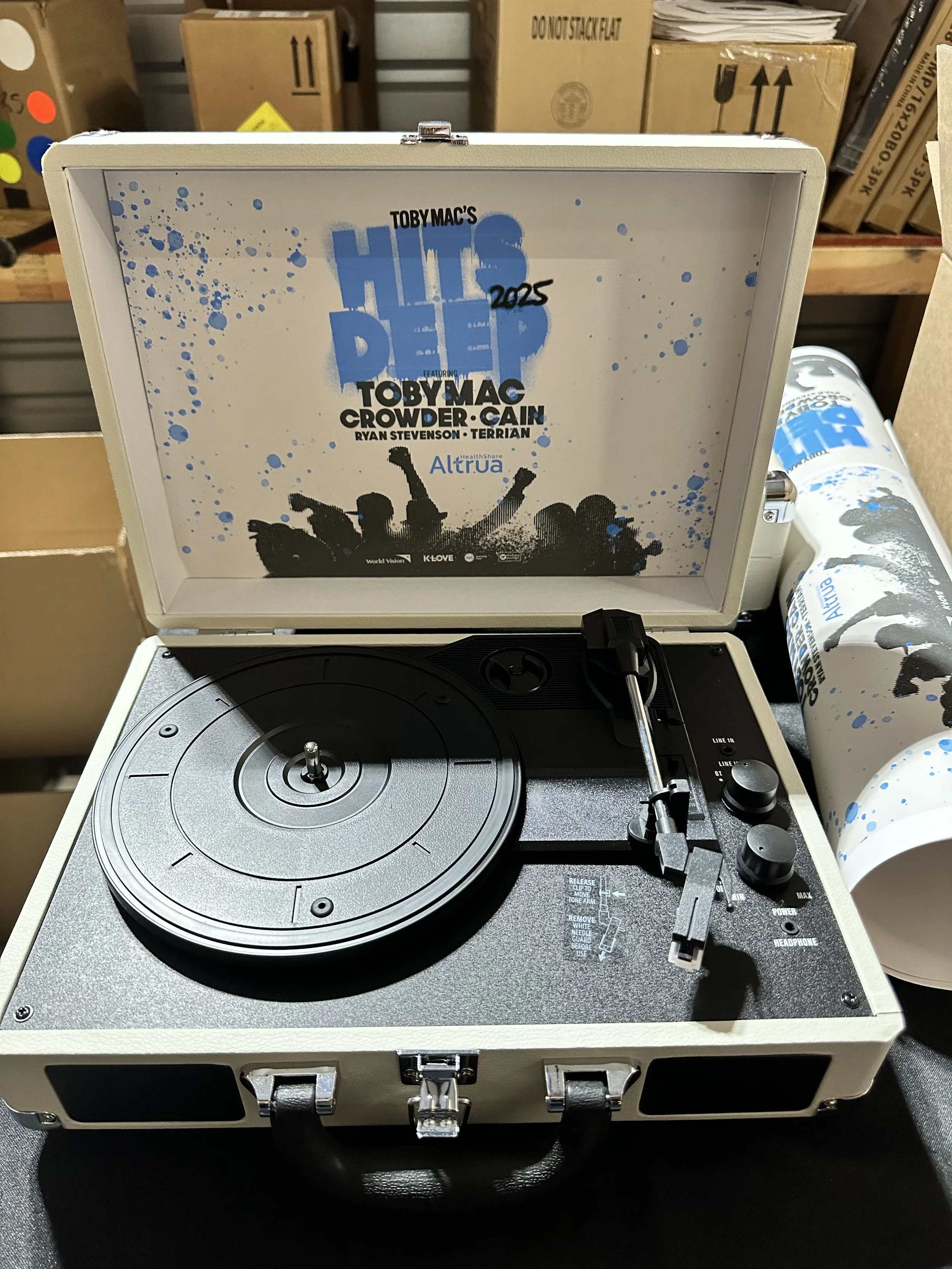 Turntable inside a white protective case with a slogan on the inside lid, featuring a black silhouette of people celebrating and blue paint splatters. The turntable is black with silver accents, with controls and a tonearm on the right side. In the background, there are cardboard boxes and a plastic bottle.