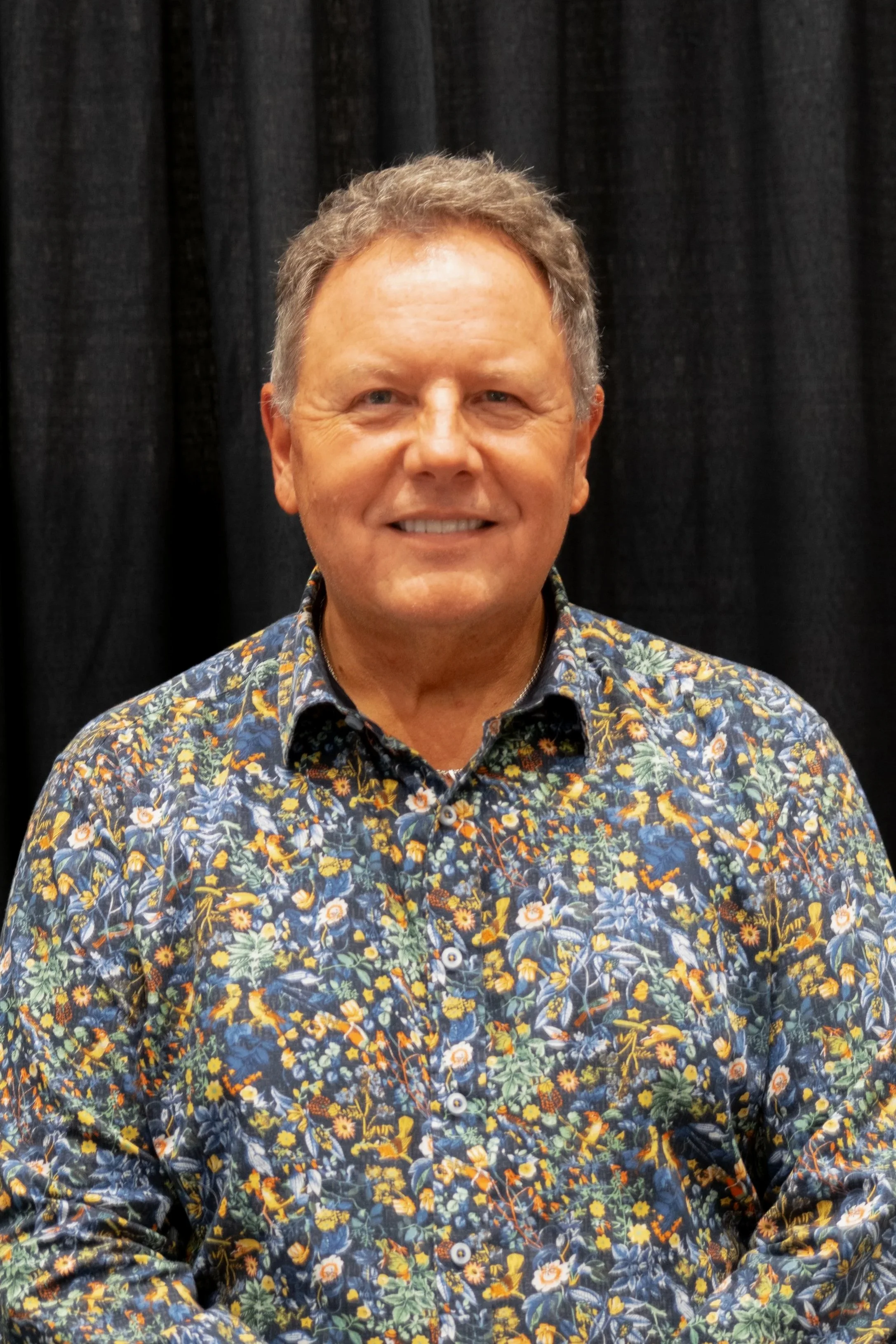 A middle-aged man with curly hair and a friendly smile, dressed in a colorful floral shirt, standing in front of a black curtain.