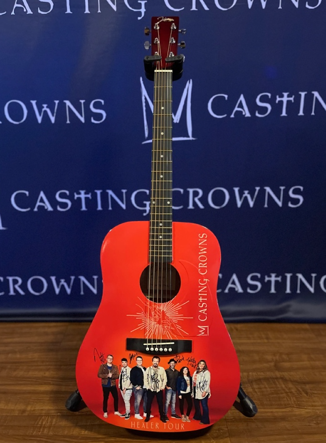 A red acoustic guitar with a photo of a band of seven autographs at the bottom, standing on a wooden surface with a blue backdrop that has "Casting Crowns" written repeatedly.