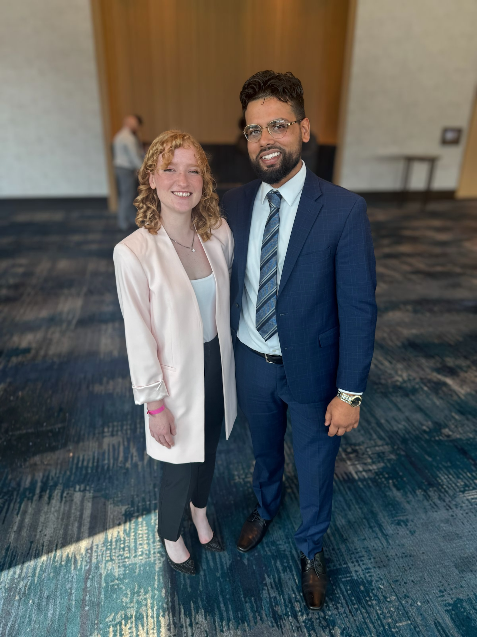 A woman and a man sitting at a wooden table in a library or bookstore. The woman is smiling and has dark hair, wearing a black blazer and checkered pants. The man is smiling, wearing a dark suit and a patterned tie. They have smartphones on the table in front of them, and there are bookshelves and a wooden partition in the background.