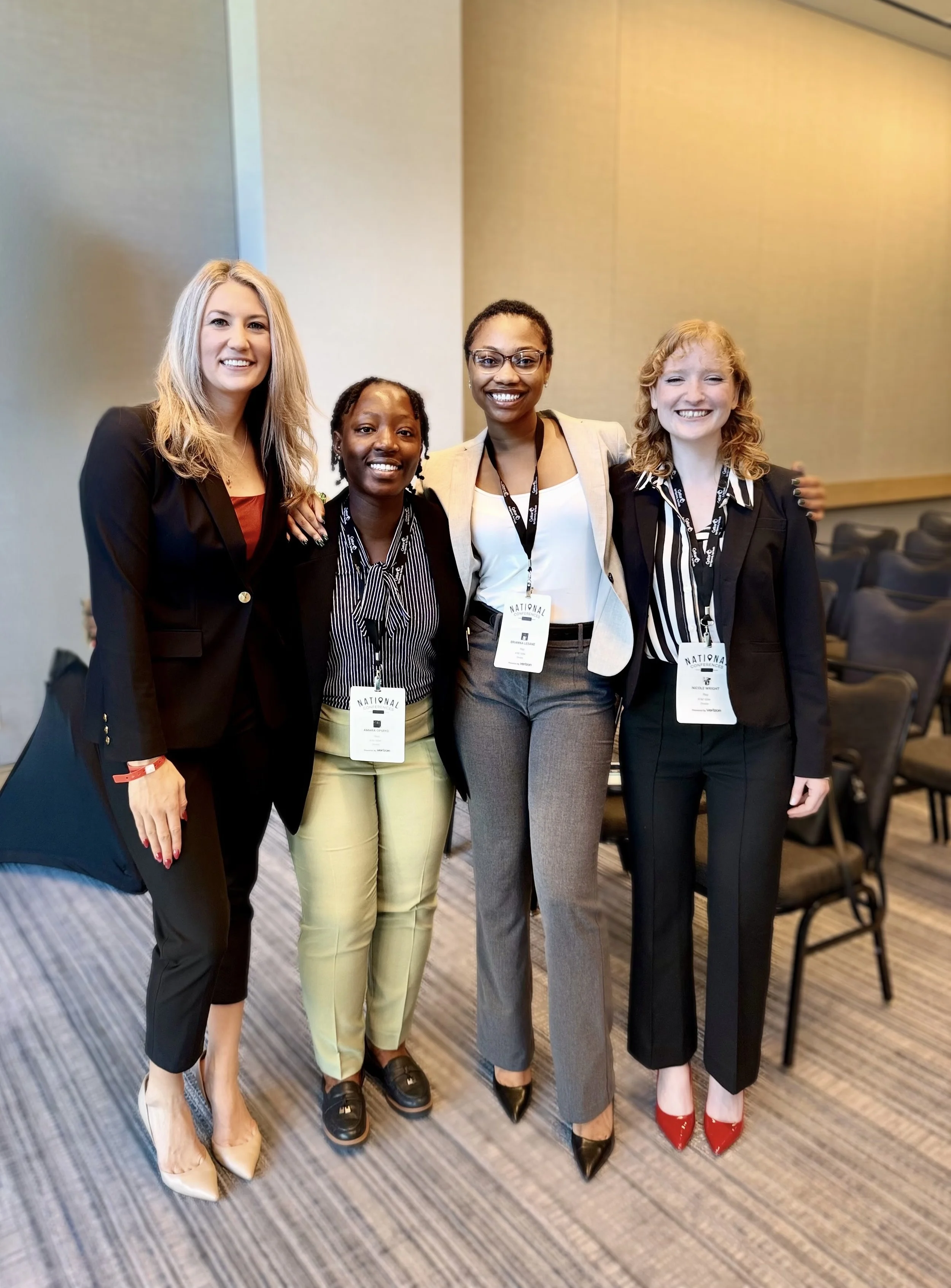 Four women standing together at a professional conference, smiling and wearing business attire with conference badges around their necks.