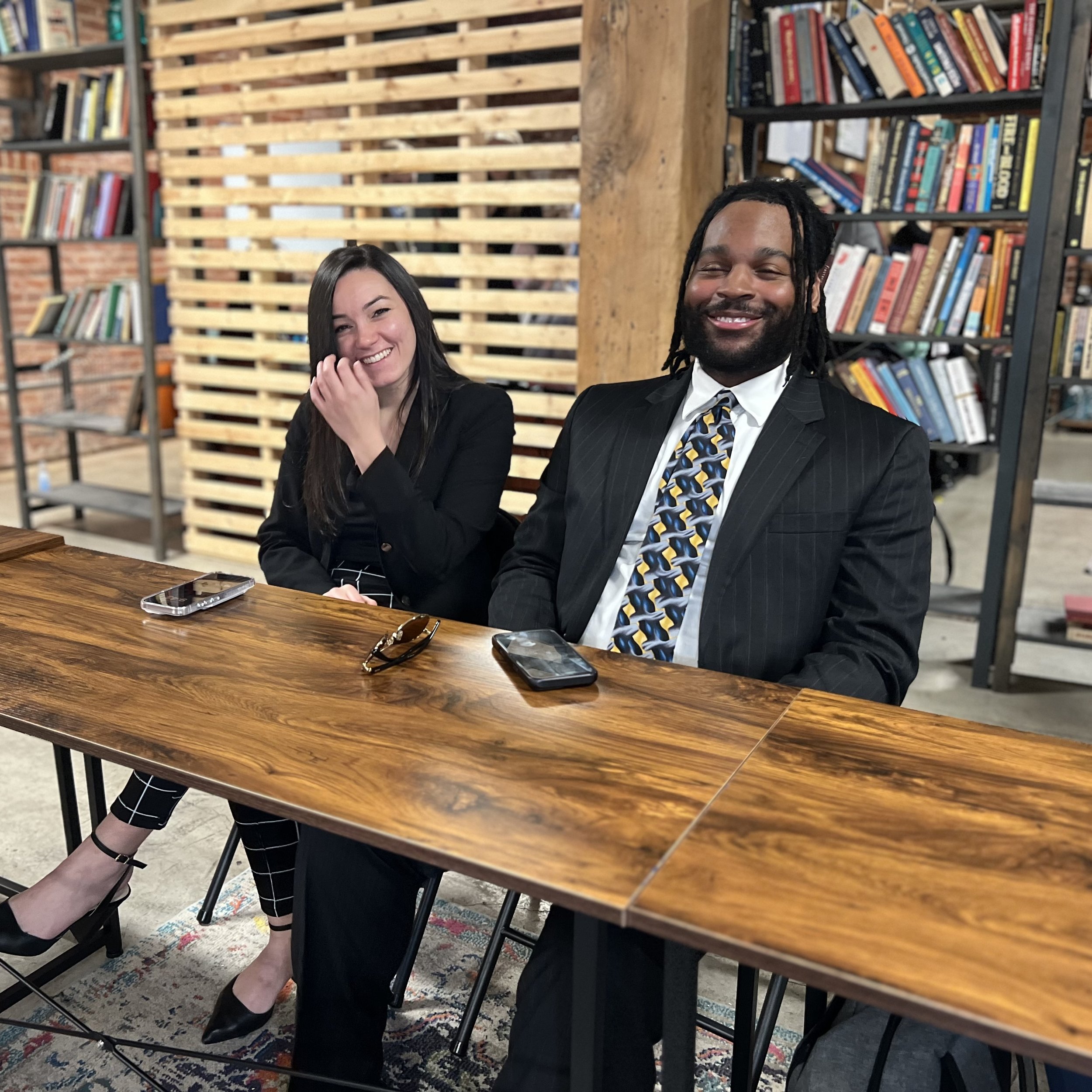 Two men are having a conversation in an indoor space with industrial decor. One man, with long dreadlocks and wearing a gray jacket, is facing the other man, who has short dark hair, dressed in a light blue blazer, black shirt, and brown shoes.