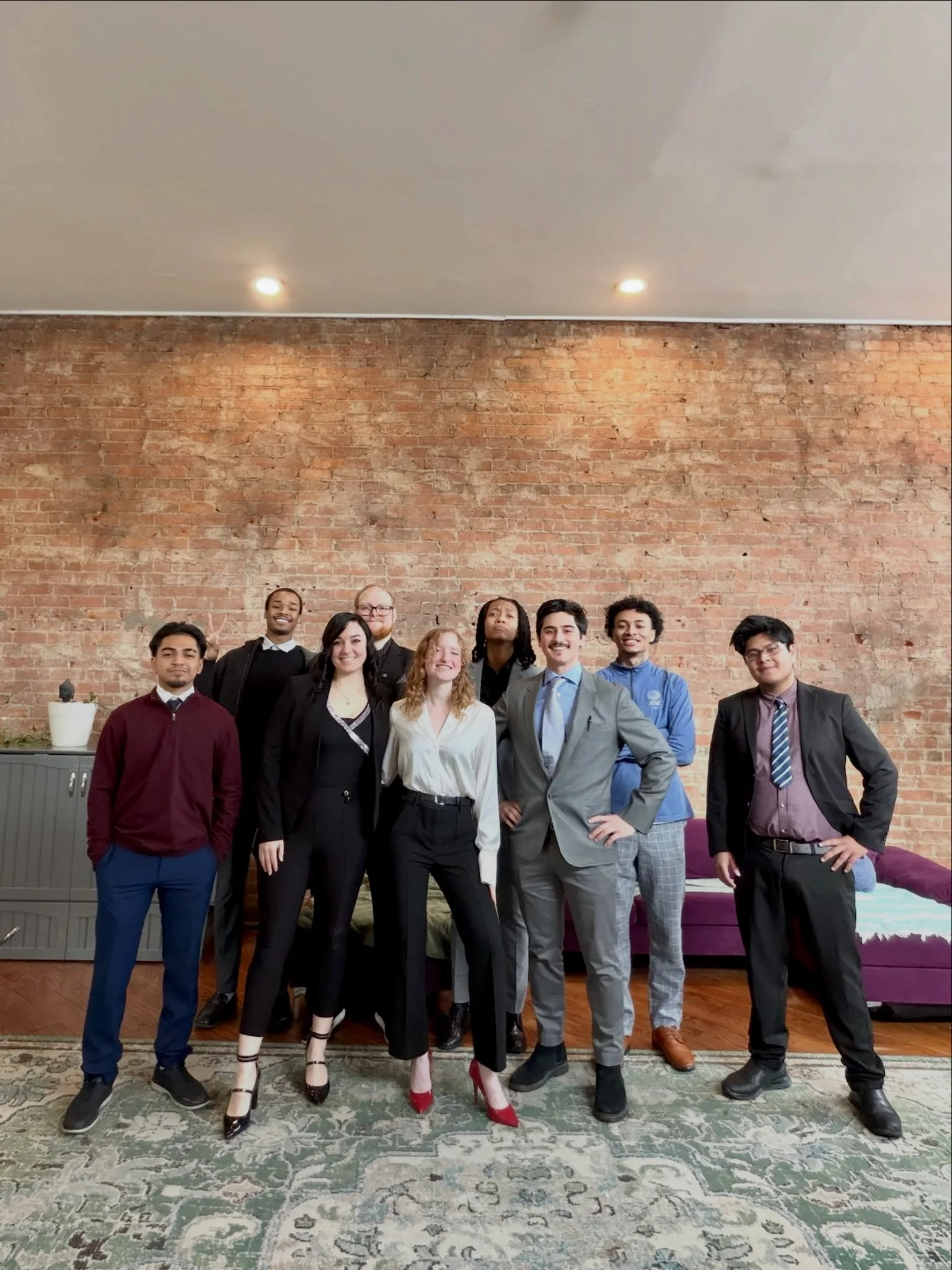 A business meeting or workshop taking place in a conference room with seven people seated around a large wooden table, a man standing near a screen giving a presentation, gift bags on the table, and laptops open in front of some attendees.