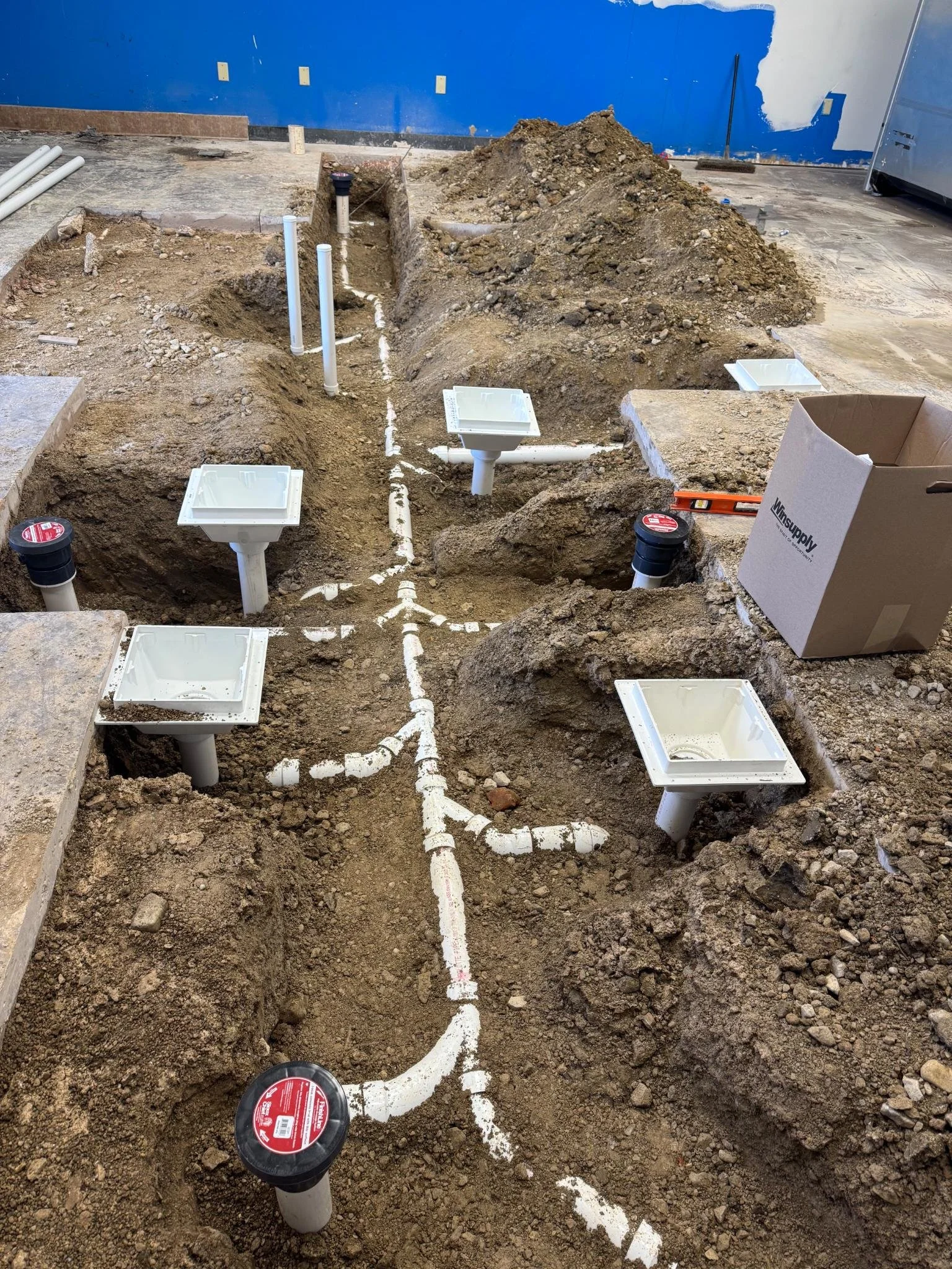 Underground plumbing pipes installation in a construction site with trenches, white PVC pipes, and a blue wall in the background.